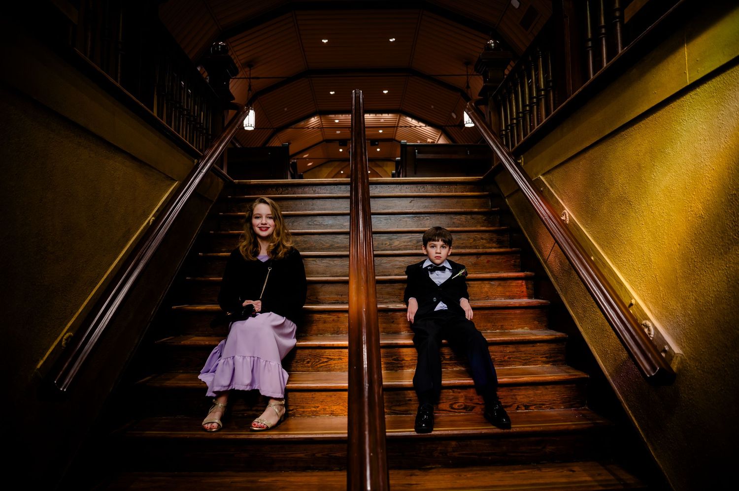 Two children sit on opposite sides of a grand wooden staircase in an elegant dimly lit venue.