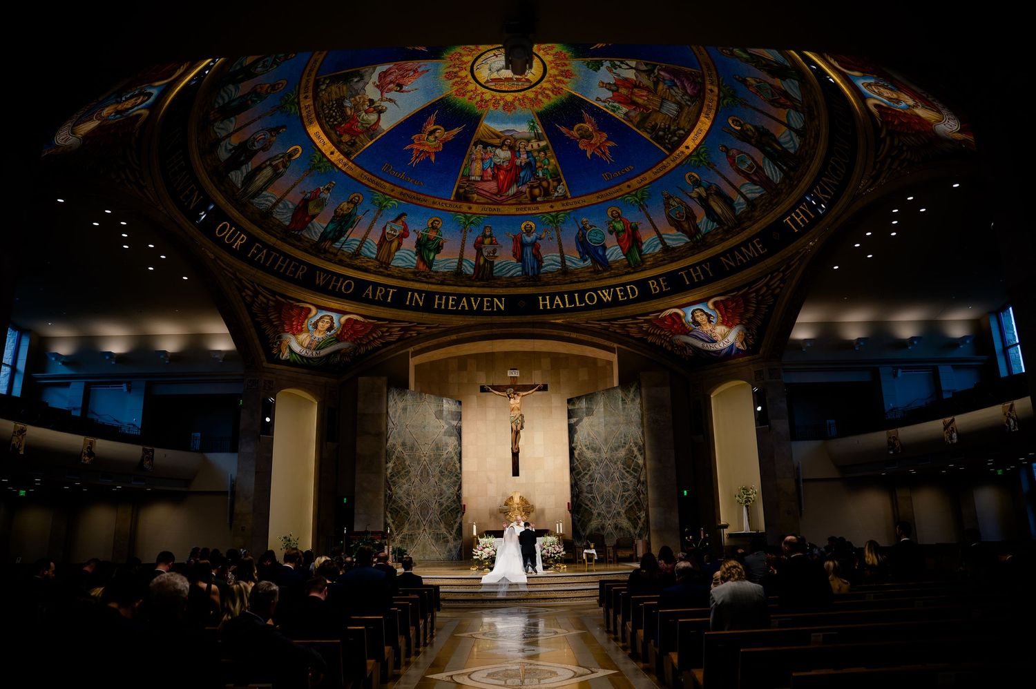Elegant Catholic church wedding ceremony under ornate painted dome with crucifix altar and seated guests in wooden pews.