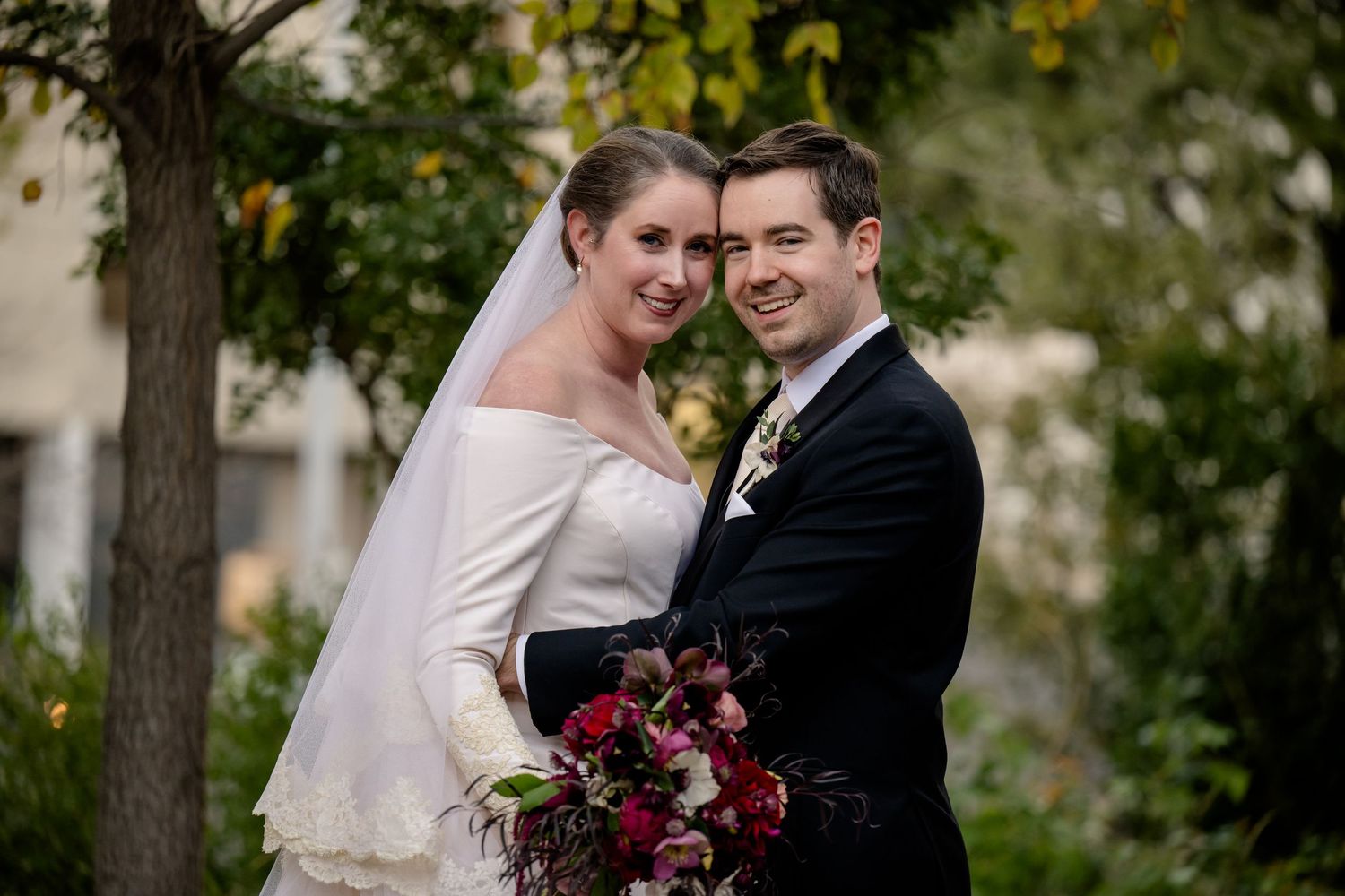 Bride in white off-shoulder gown with veil and groom in black suit pose together outdoors holding a dark floral bouquet.