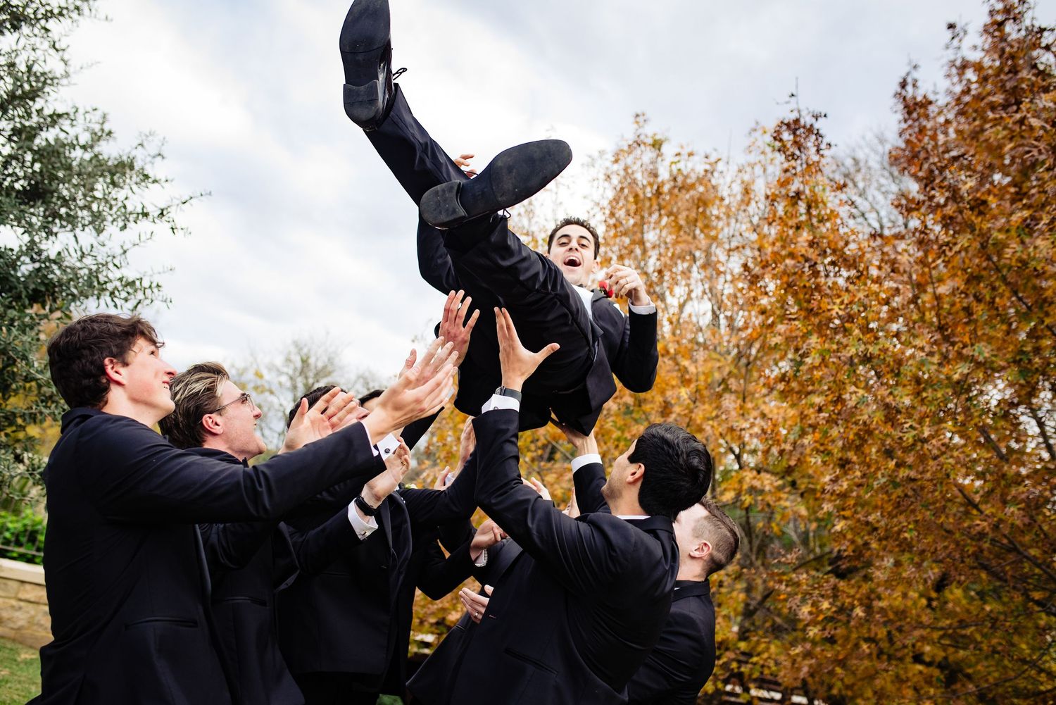 Groomsmen in black suits tossing groom into the air outdoors with colorful autumn trees in the background.