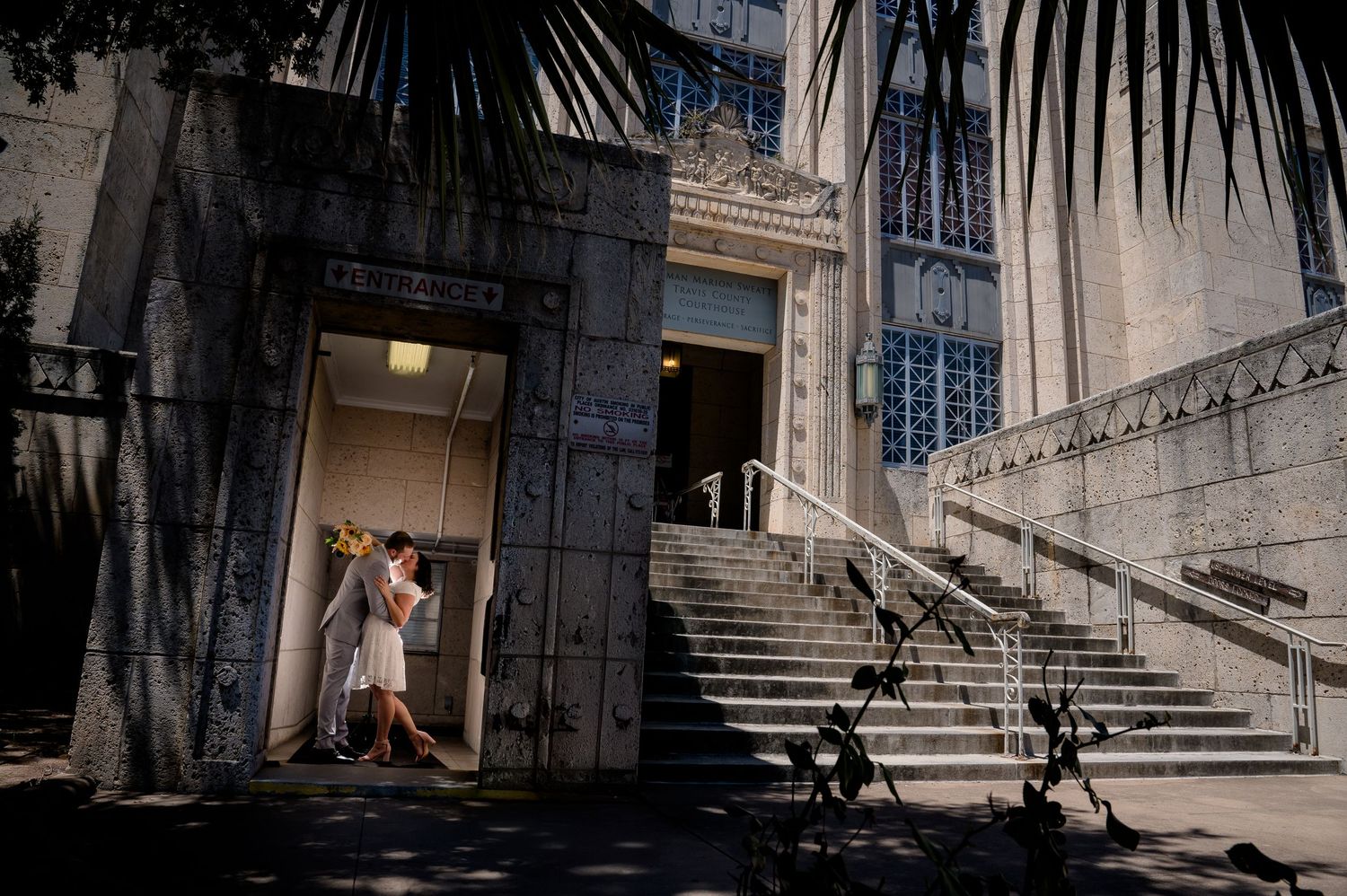 Couple sharing a romantic kiss in the shadowed doorway of an ornate stone building with grand stairs.