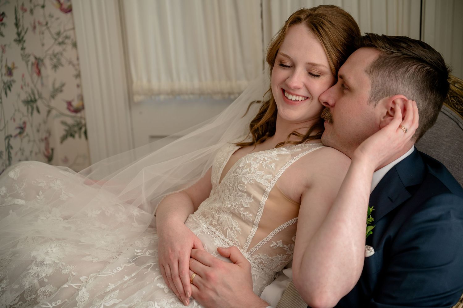 Bride in lace gown smiles warmly as groom in navy suit embraces her tenderly during intimate wedding portrait.