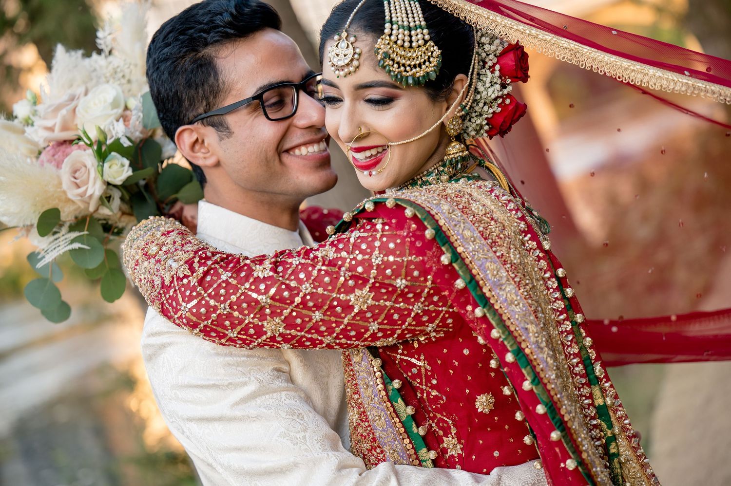 A joyful South Asian couple in traditional wedding attire, bride in red lehenga with gold jewelry embracing groom in white sherwani.