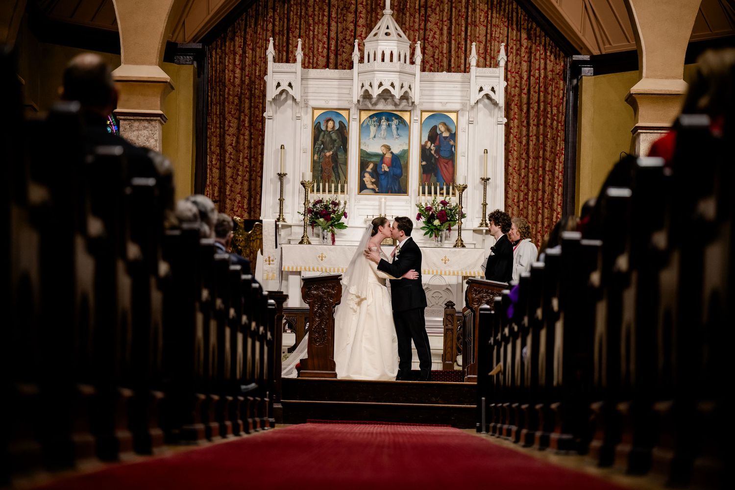 Bride and groom share first kiss at church altar during elegant wedding ceremony with red carpet aisle.