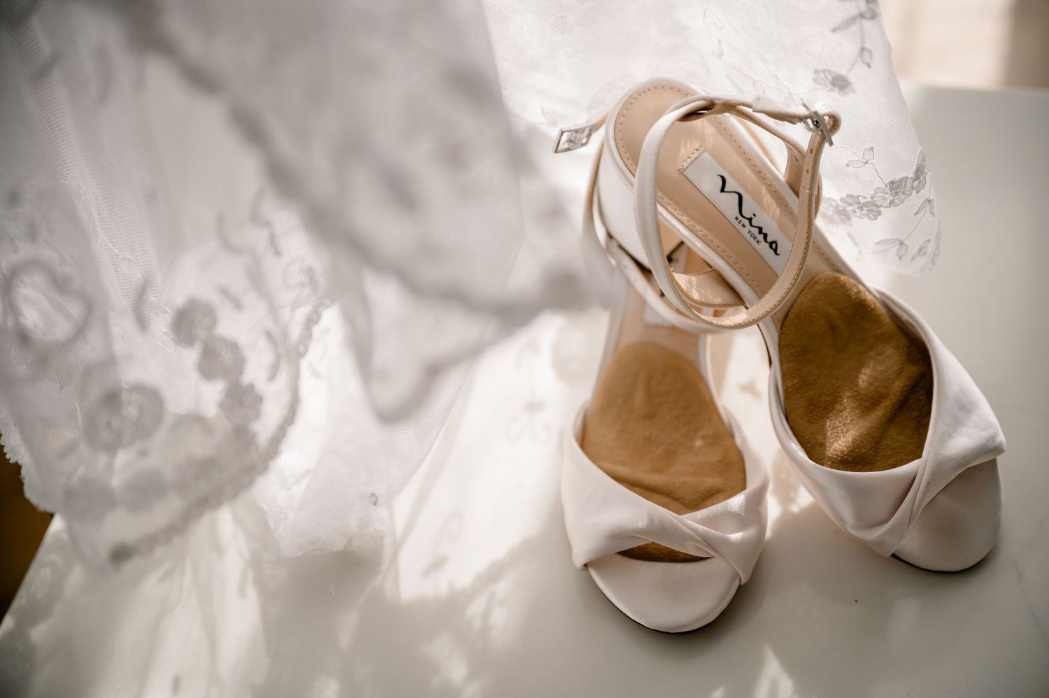 Elegant white bridal heels placed beside a lace wedding dress.