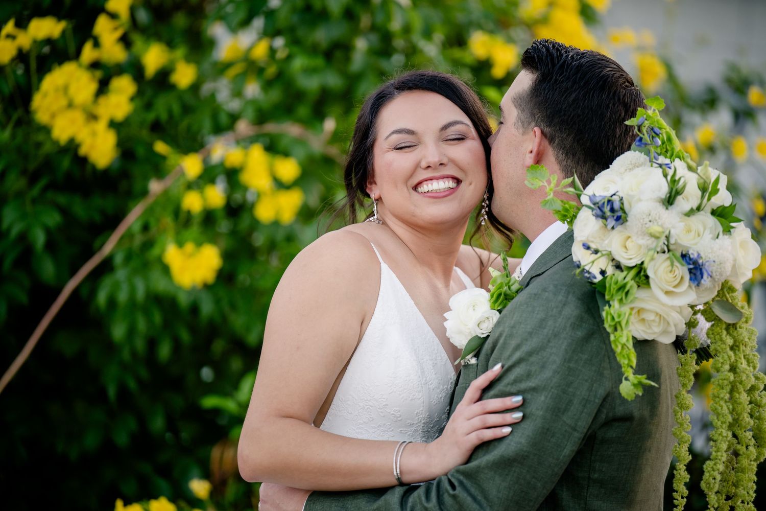 A joyful bride laughs as her groom kisses her cheek, holding a white floral bouquet with yellow blooms in the background.