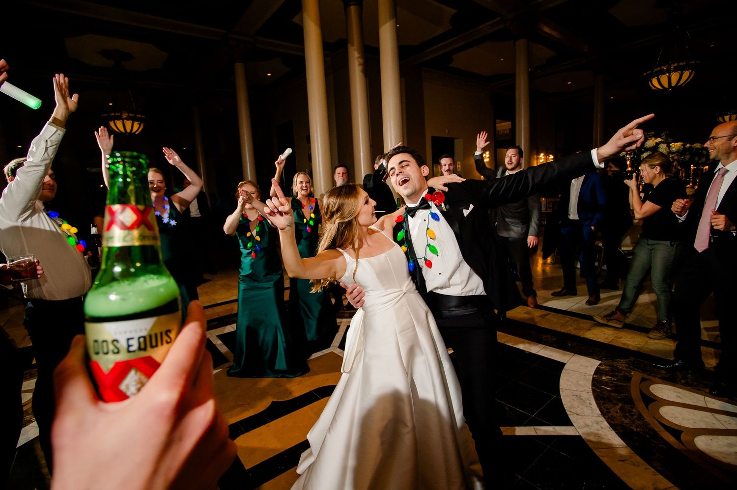 Bride and groom dancing joyfully at wedding reception with guests celebrating and green beer bottle in foreground.