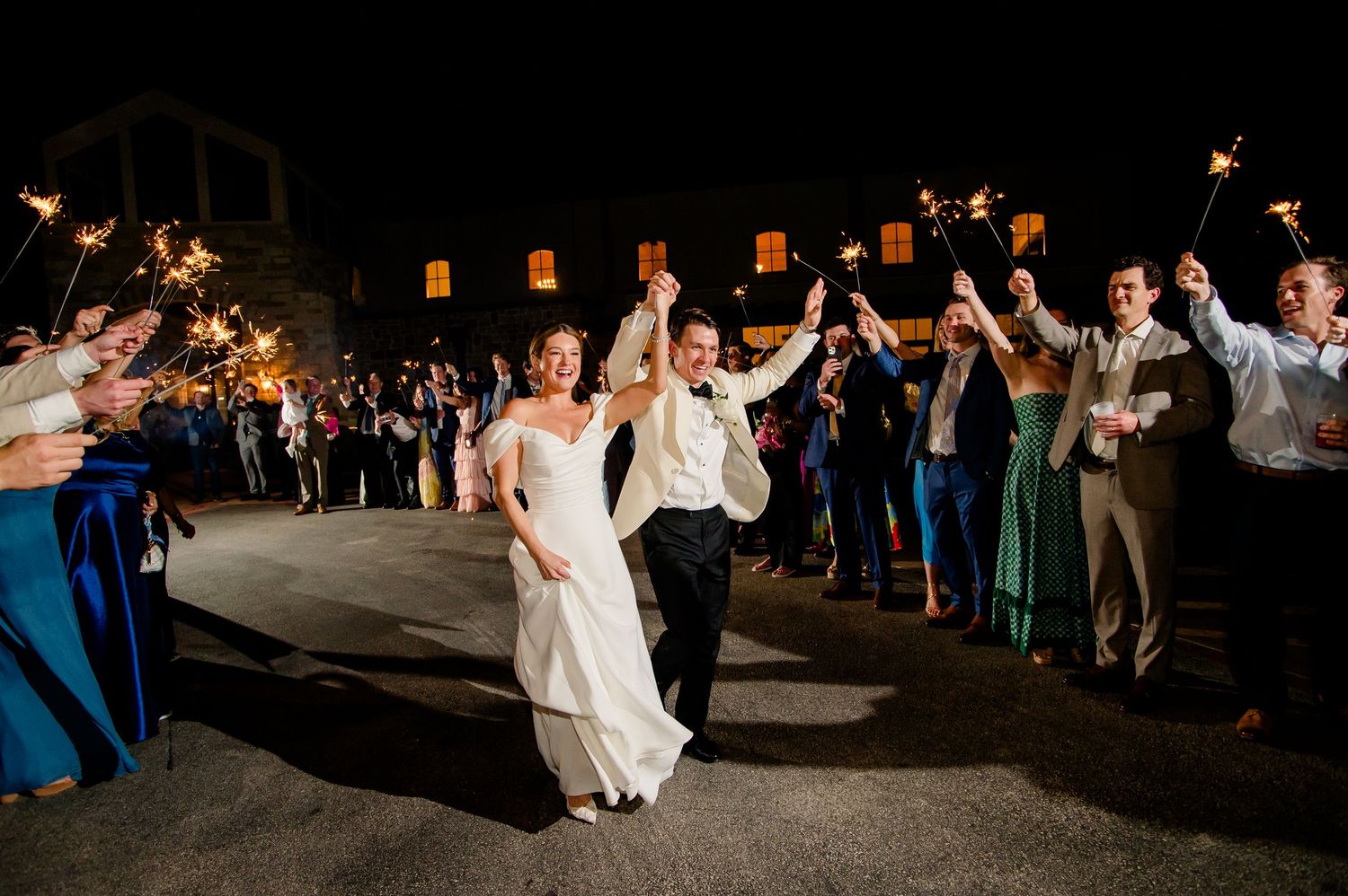 Bride and groom exit wedding reception through sparkler tunnel surrounded by cheering guests at night.