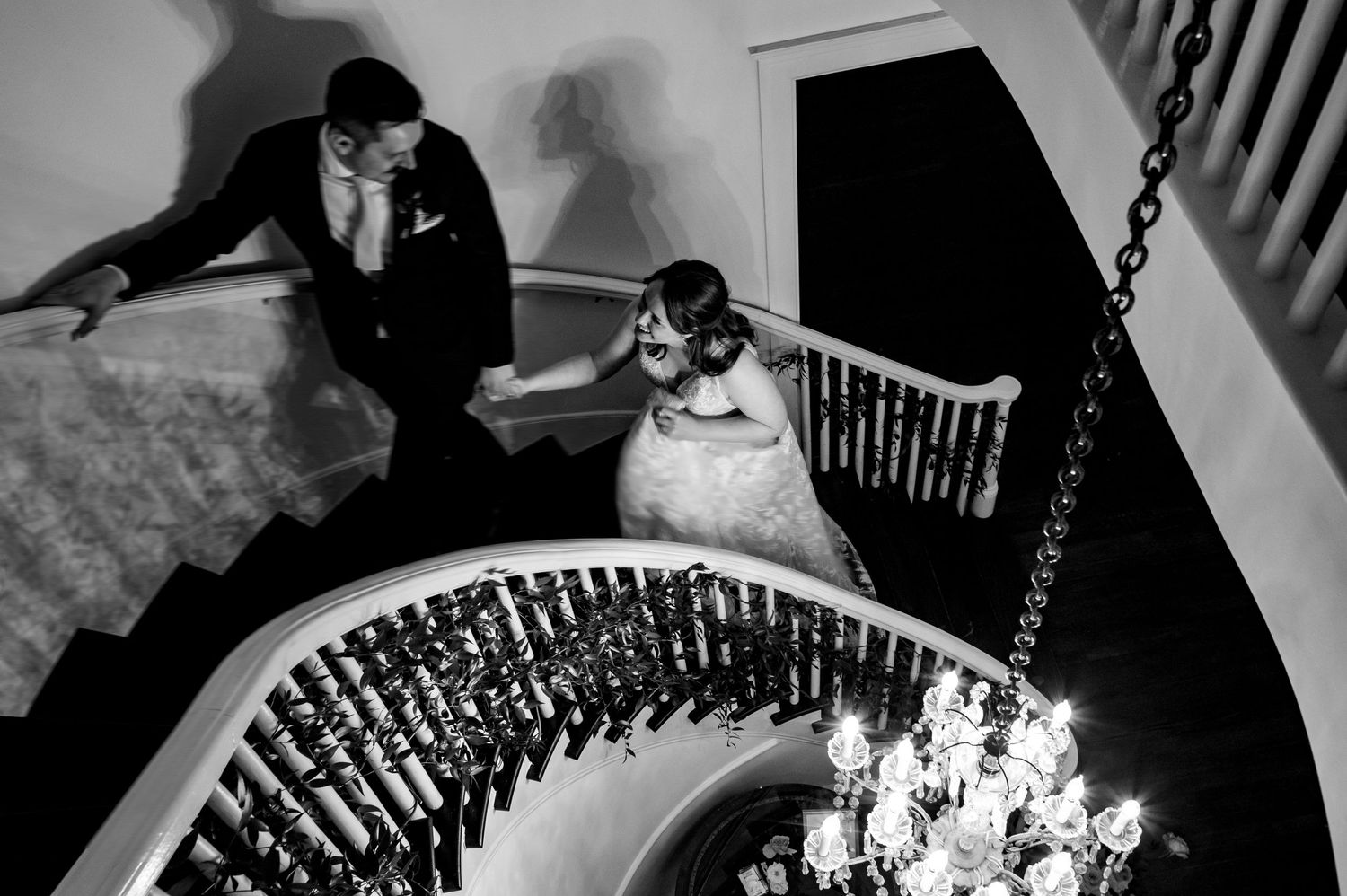 Black and white wedding photo of bride and groom on elegant curved staircase with chandelier below.
