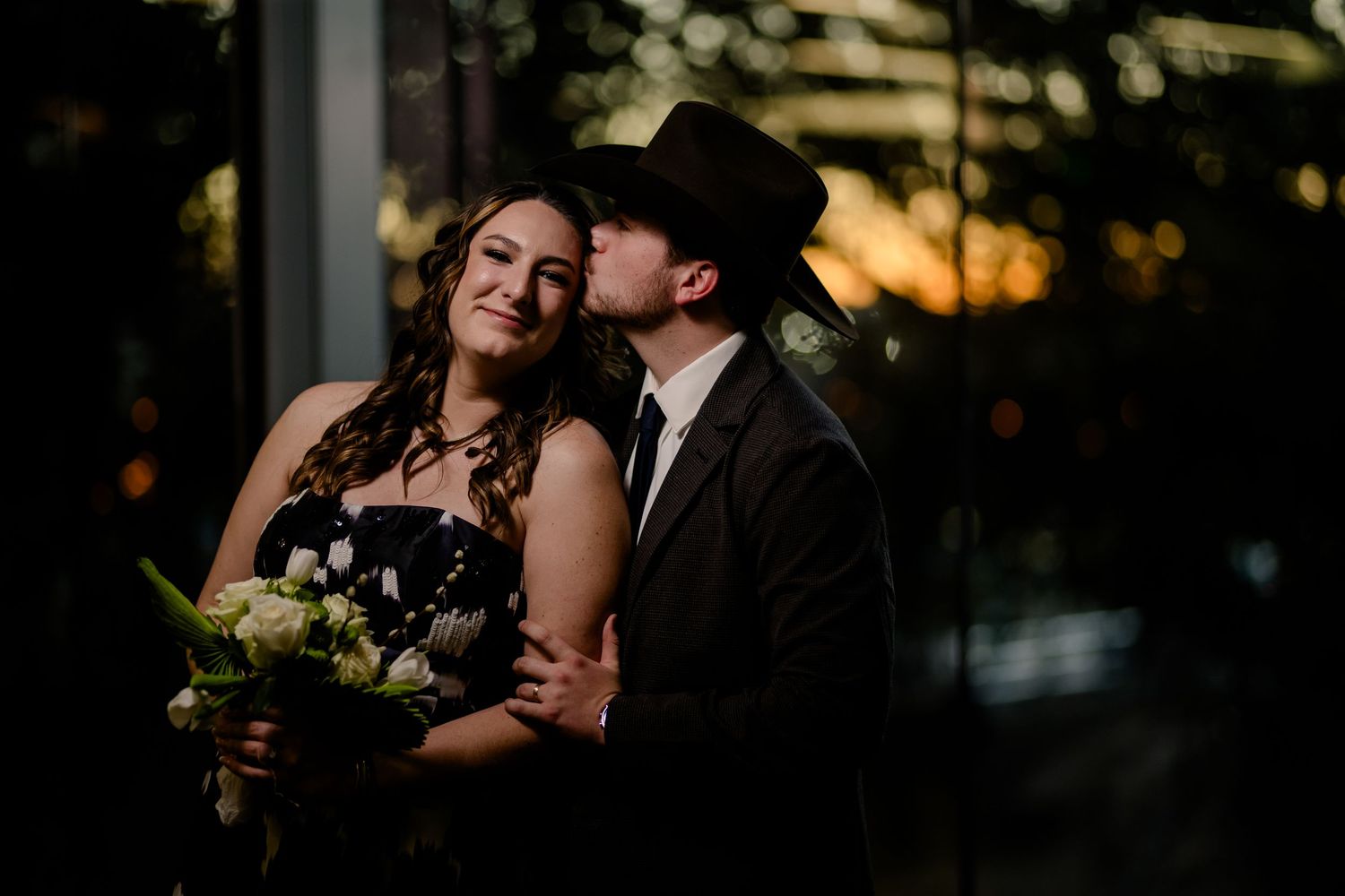 Couple in formal wear at sunset, groom in black cowboy hat kissing bride holding white floral bouquet.