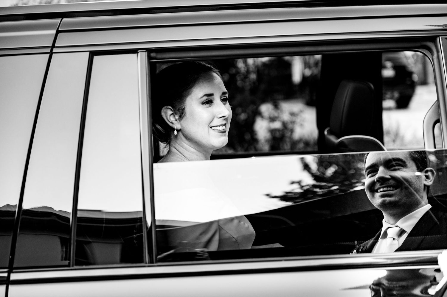 Black and white photo of a smiling bride inside a car, with a man in a suit visible through the window reflection.