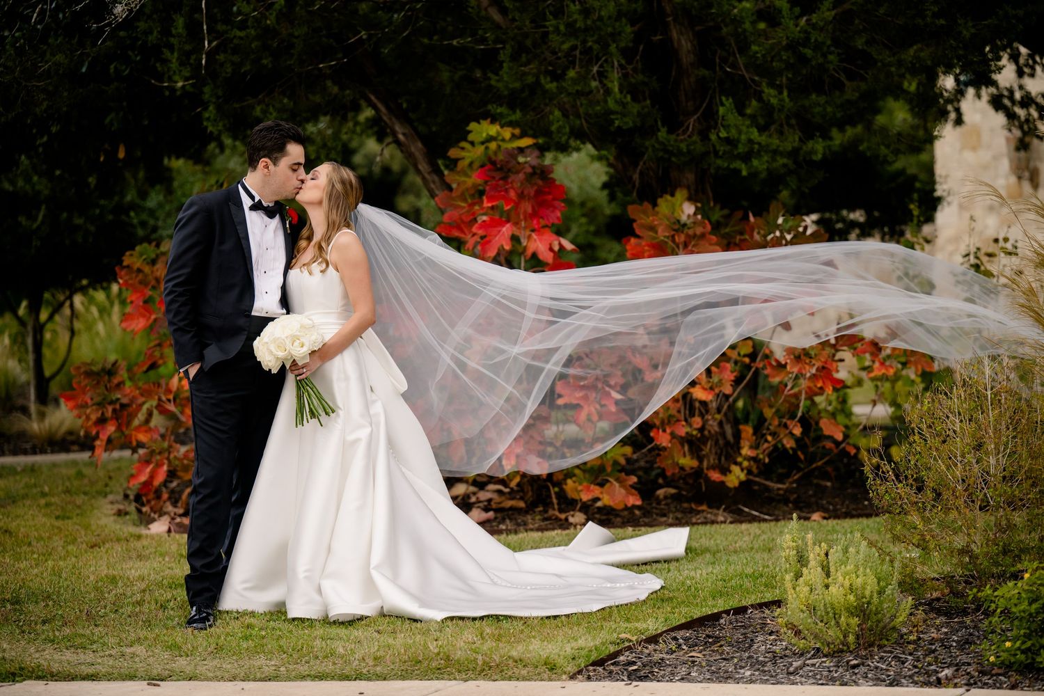 Bride and groom kiss outdoors as long veil flows in wind with colorful autumn foliage behind them.