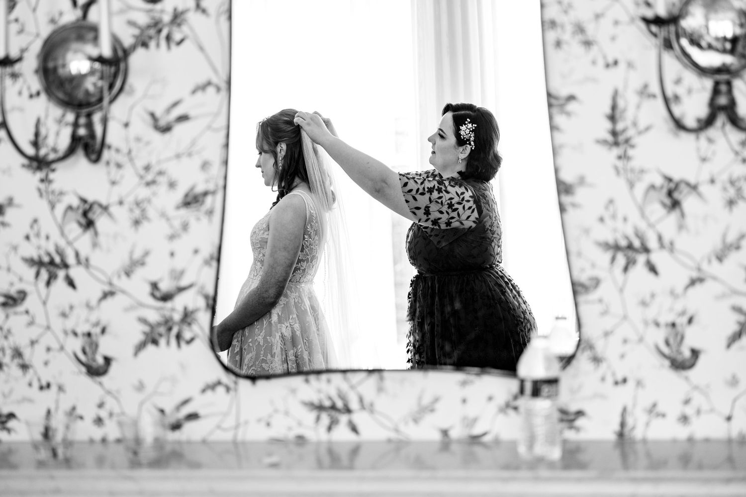 Black and white photo of a woman adjusting a bride's veil in a mirror reflection, with floral wallpaper backdrop.
