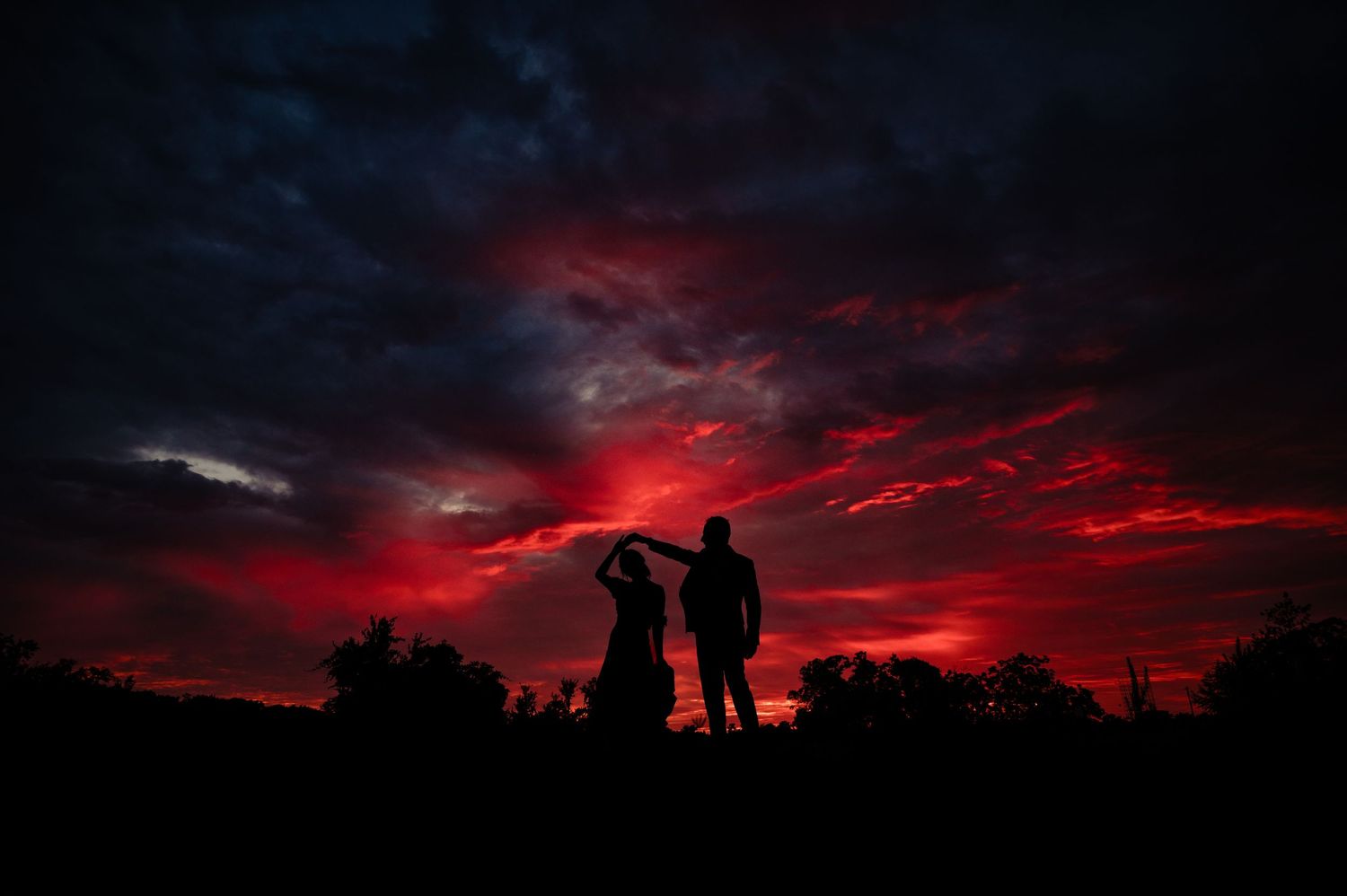 Silhouettes of a couple standing against a dramatic red and crimson sunset sky with dark storm clouds.