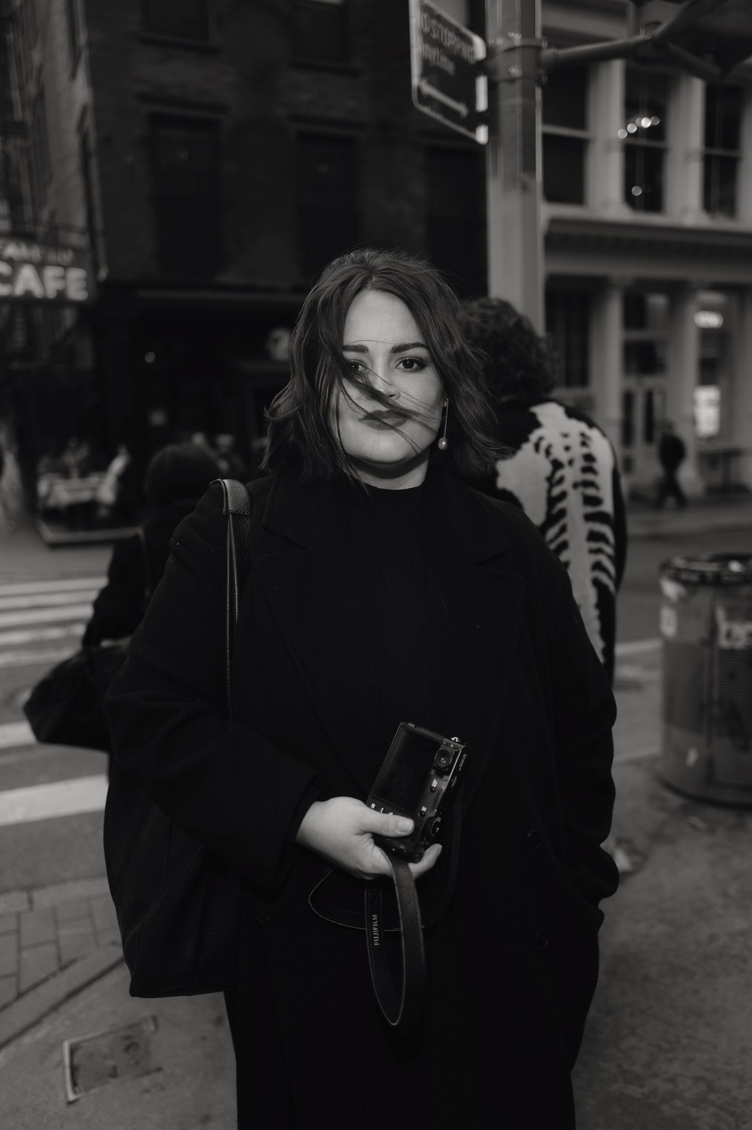Black and white street photo of a woman in a black coat holding a camera, with hair blowing across her face.