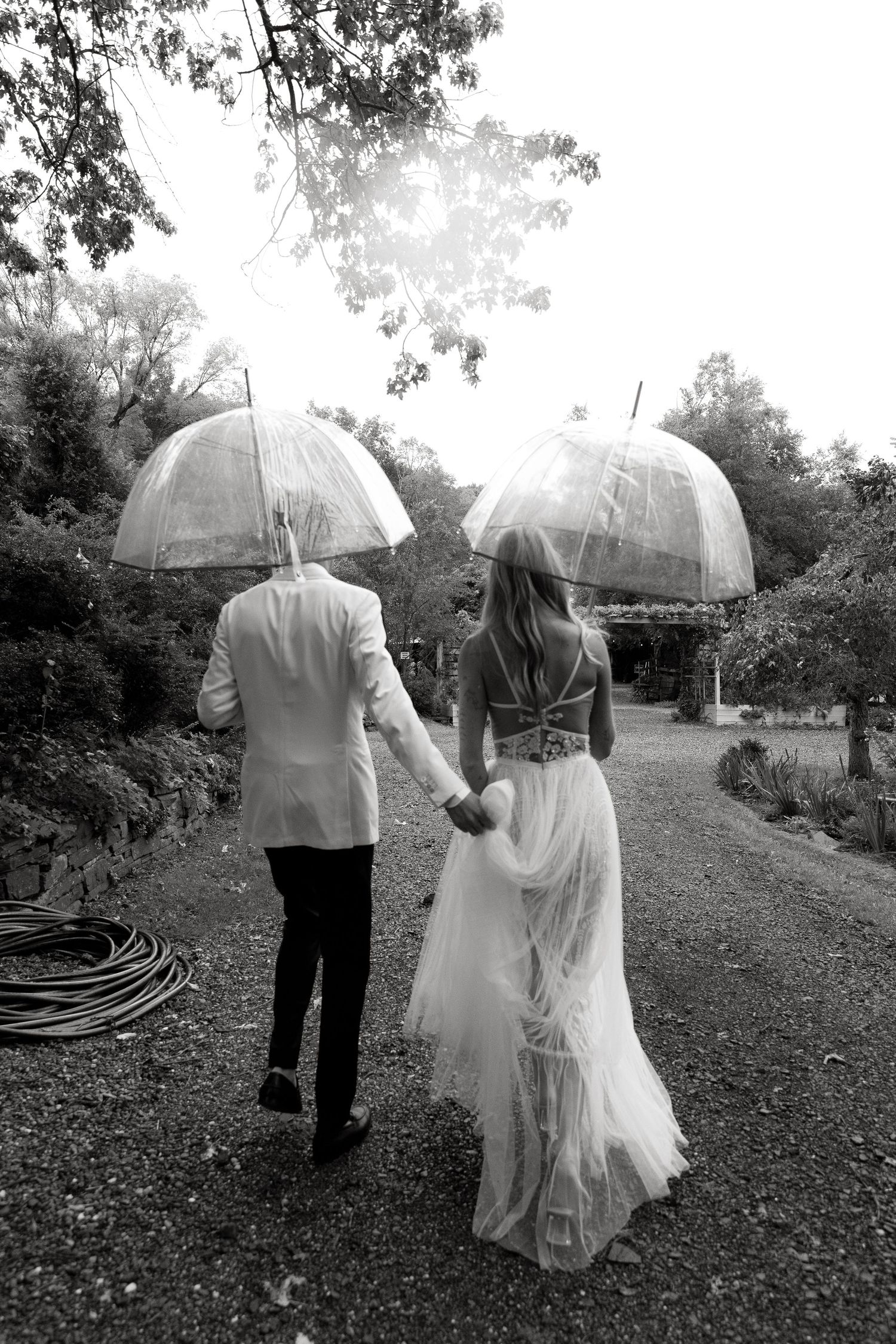 Black and white photo of a bride and groom walking hand in hand under clear dome umbrellas on a garden path.