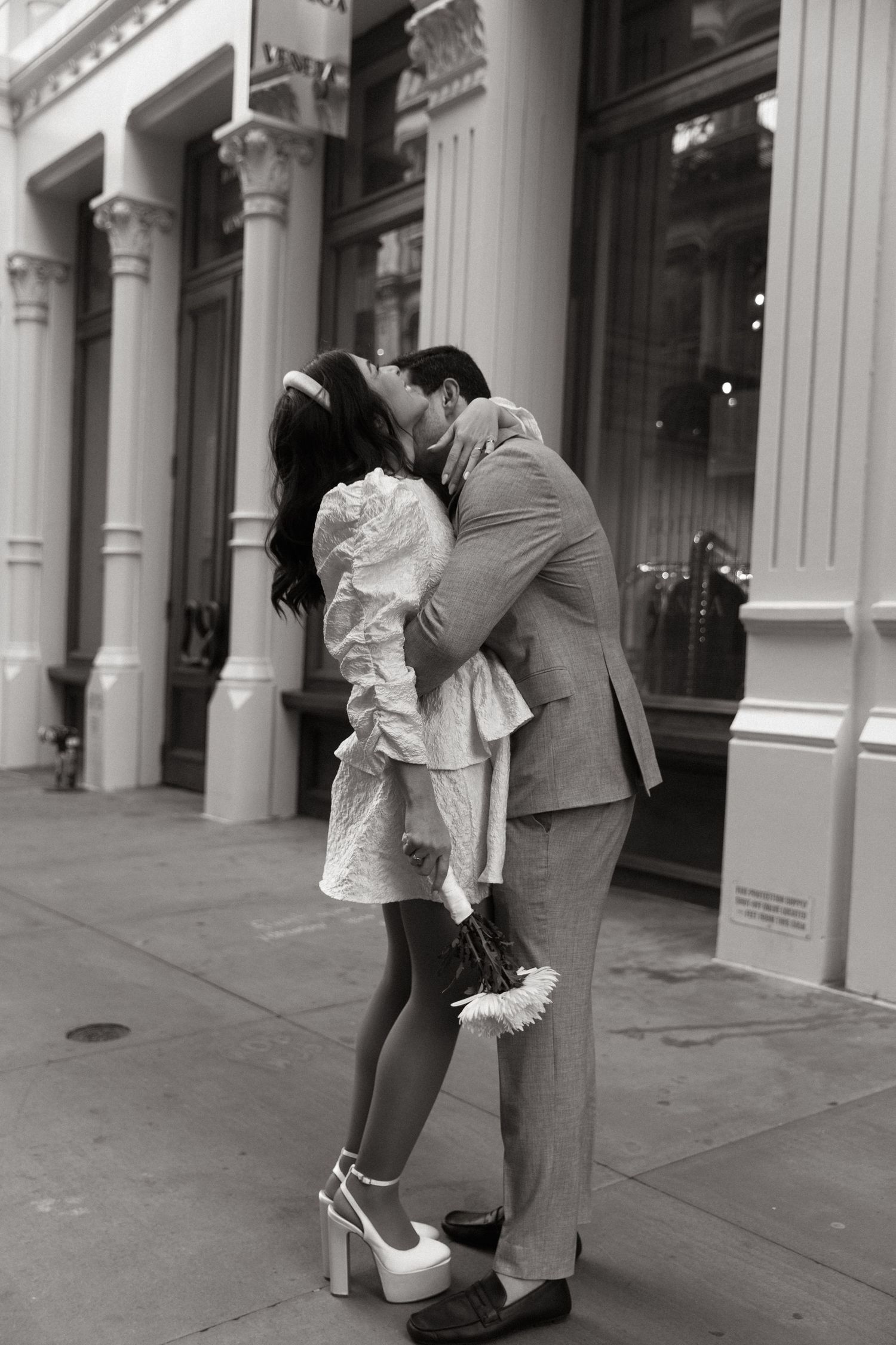 A romantic black and white photo of a couple sharing a passionate kiss in front of a building with elegant columns.