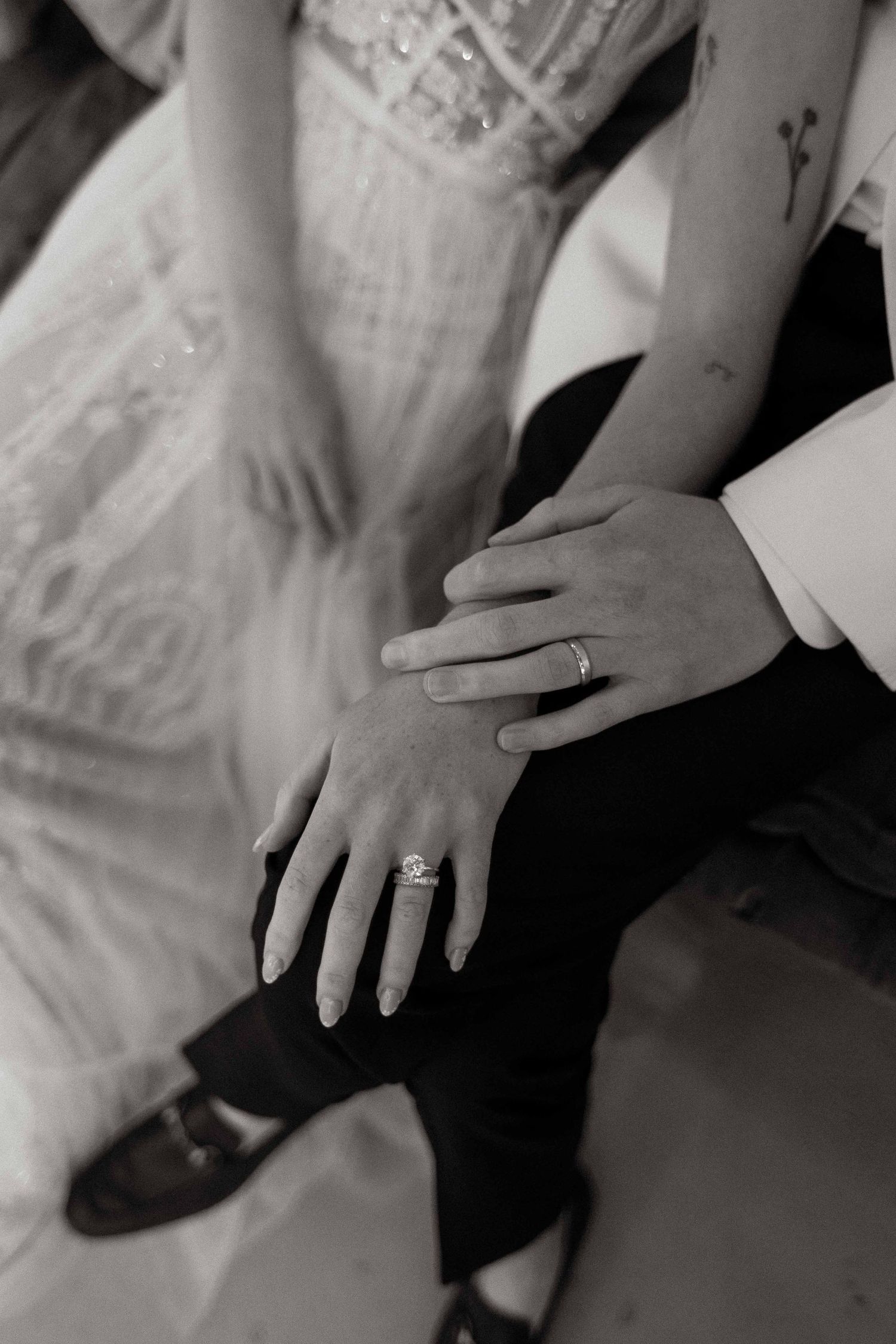 Black and white photo shows two hands with wedding rings touching, suggesting a romantic and intimate wedding moment.
