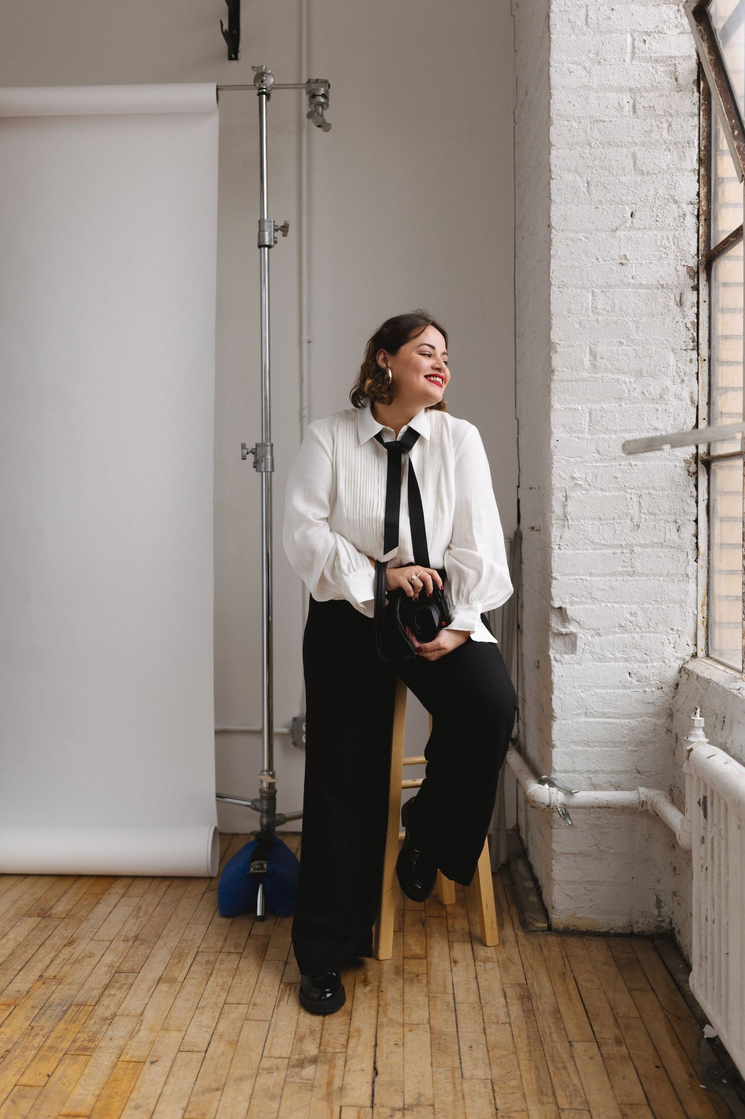 A photographer wearing a white blouse and black tie sits casually on a stool in a bright studio with white brick walls.