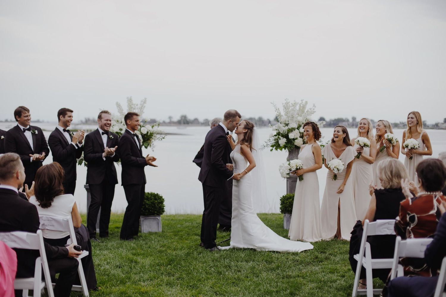 Bride and groom share first kiss at outdoor waterfront wedding ceremony with bridal party and seated guests.