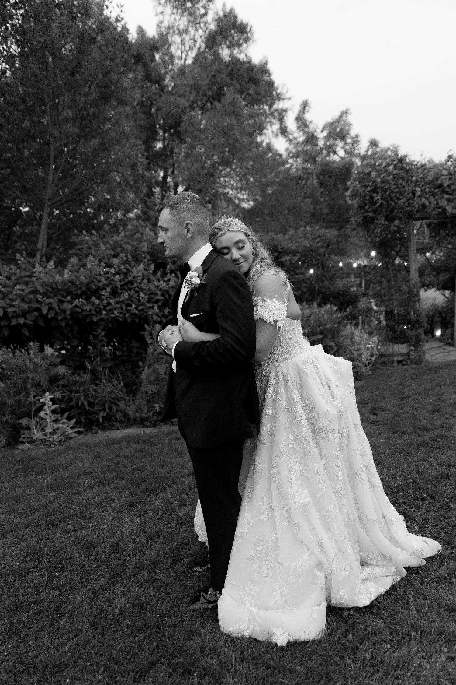 Black and white wedding photo showing couple embracing in a garden setting, with the bride in a lace gown.