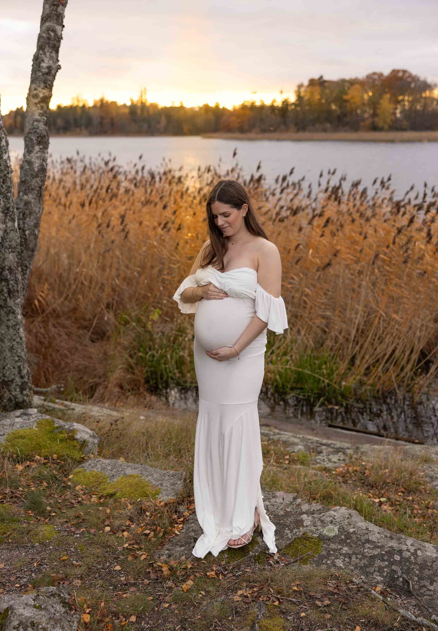 Gravidfotografering drevviken Haninge gravidkvinna håller om sin mage och har på sig en vit klänning gravidporträtt av emeraldweddings