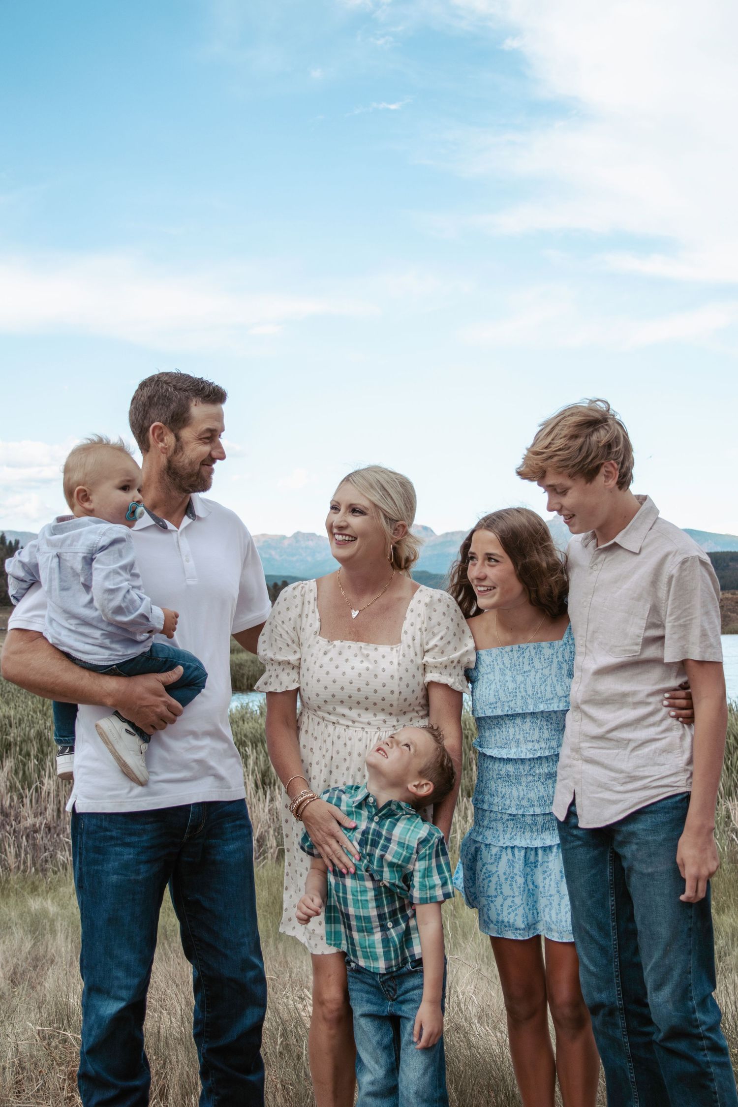 A family stands together outdoors against a blue sky background in casual clothing on a grassy hillside.