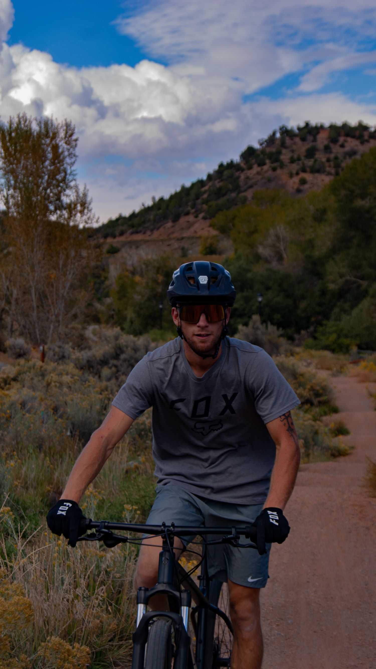 Mountain biker on dirt trail surrounded by desert brush and hills under blue sky.