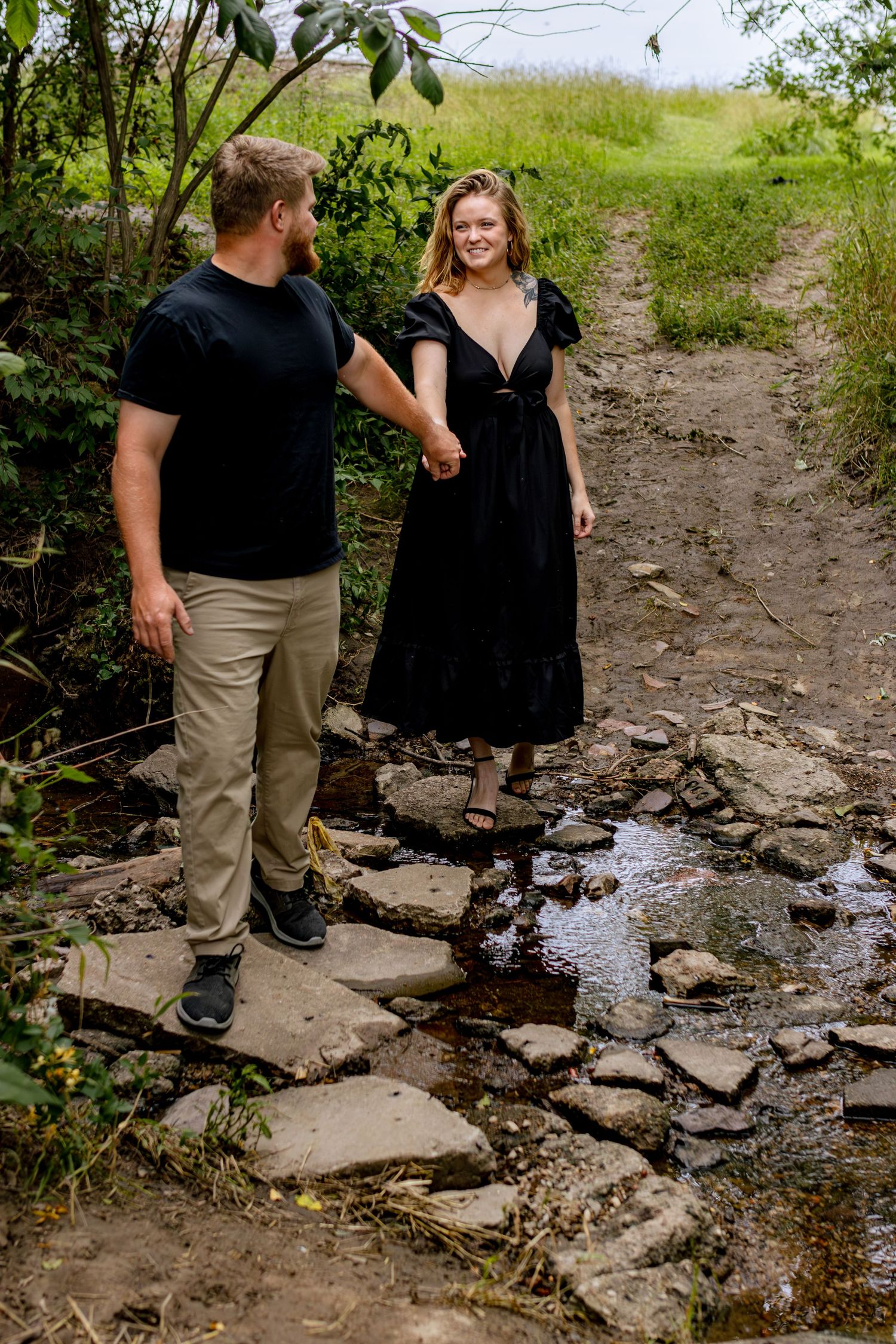 Two people walk along a rocky creek bed surrounded by green foliage on a nature trail.