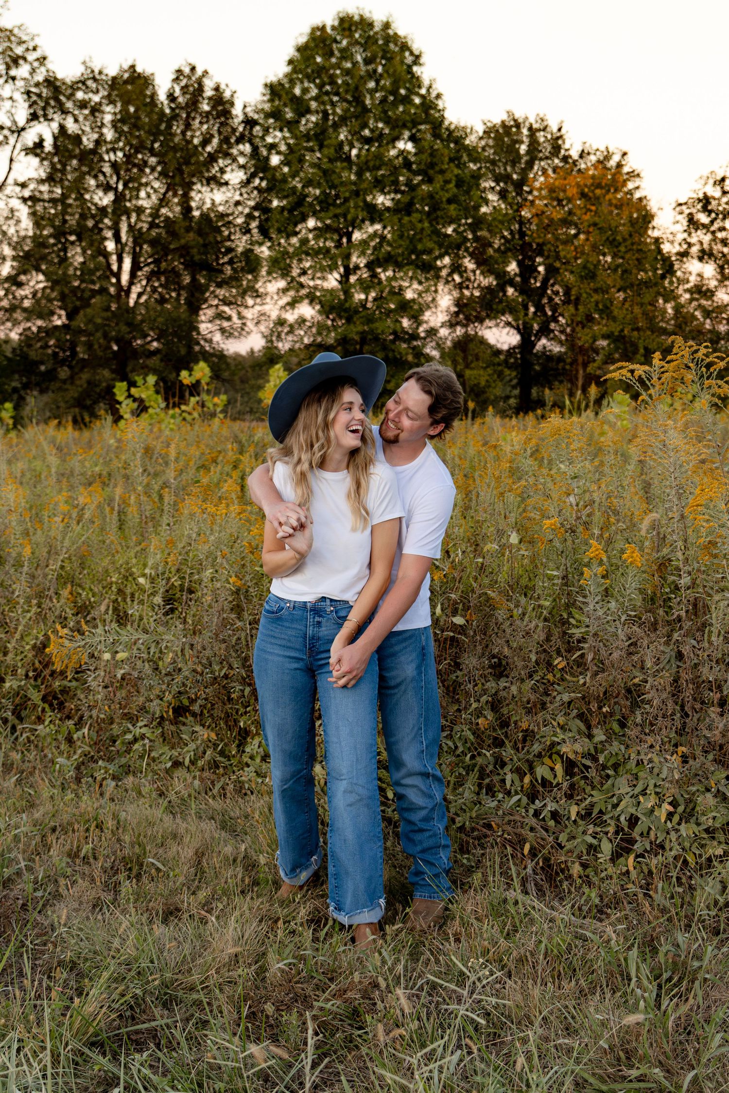 A couple stands together in a wildflower field at sunset wearing matching white shirts and blue jeans.