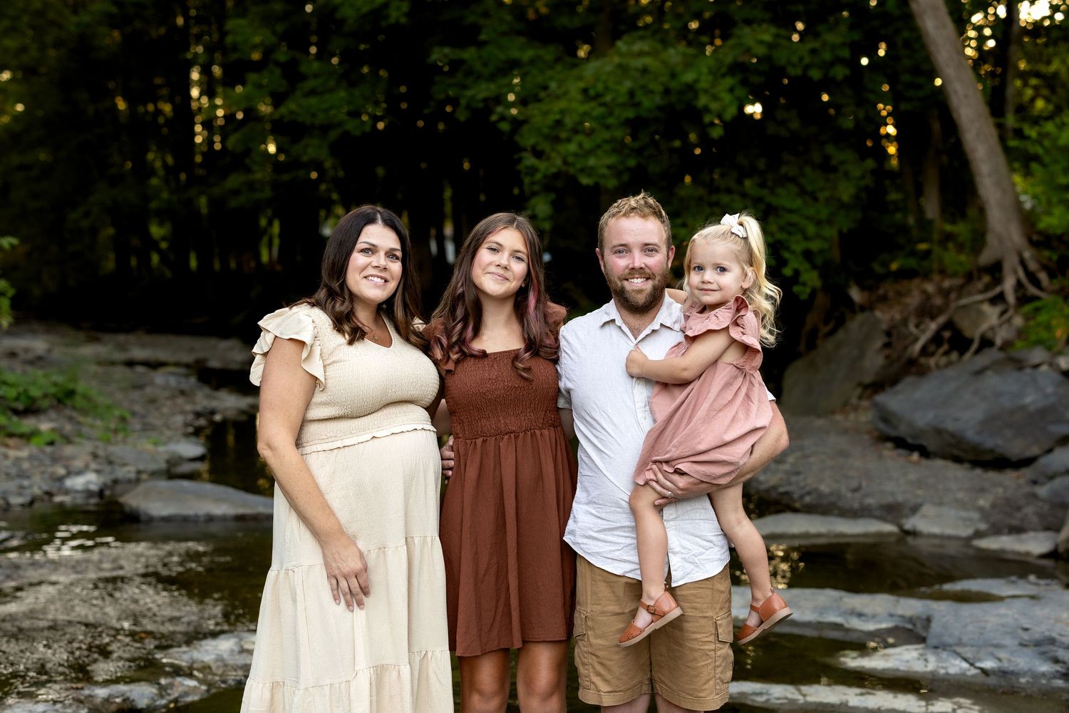 A group of people in coordinated brown and cream outfits pose together by a rocky stream in a forest setting.