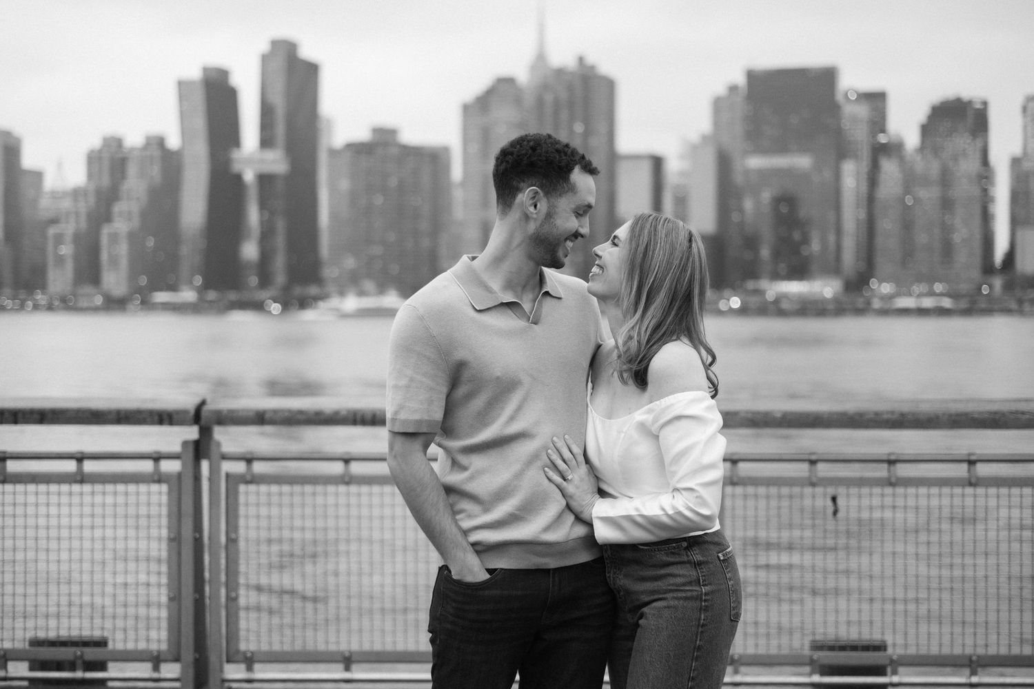 Couple embracing with NYC skyline backdrop in black and white engagement photo by the waterfront.