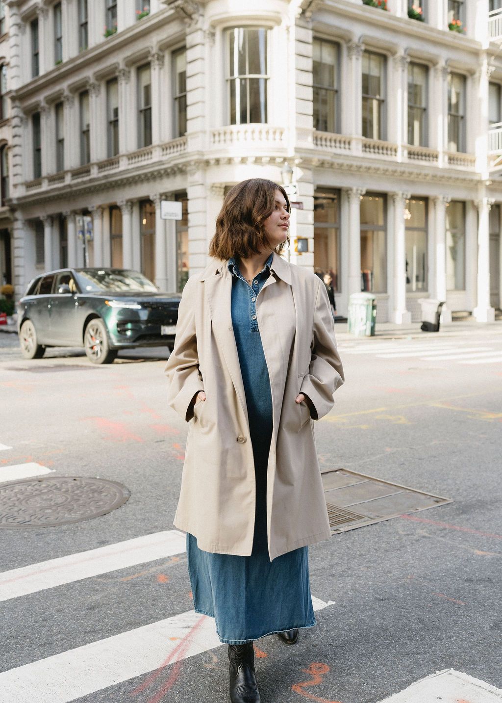 Person in beige coat and denim walking through urban street with classic architecture.