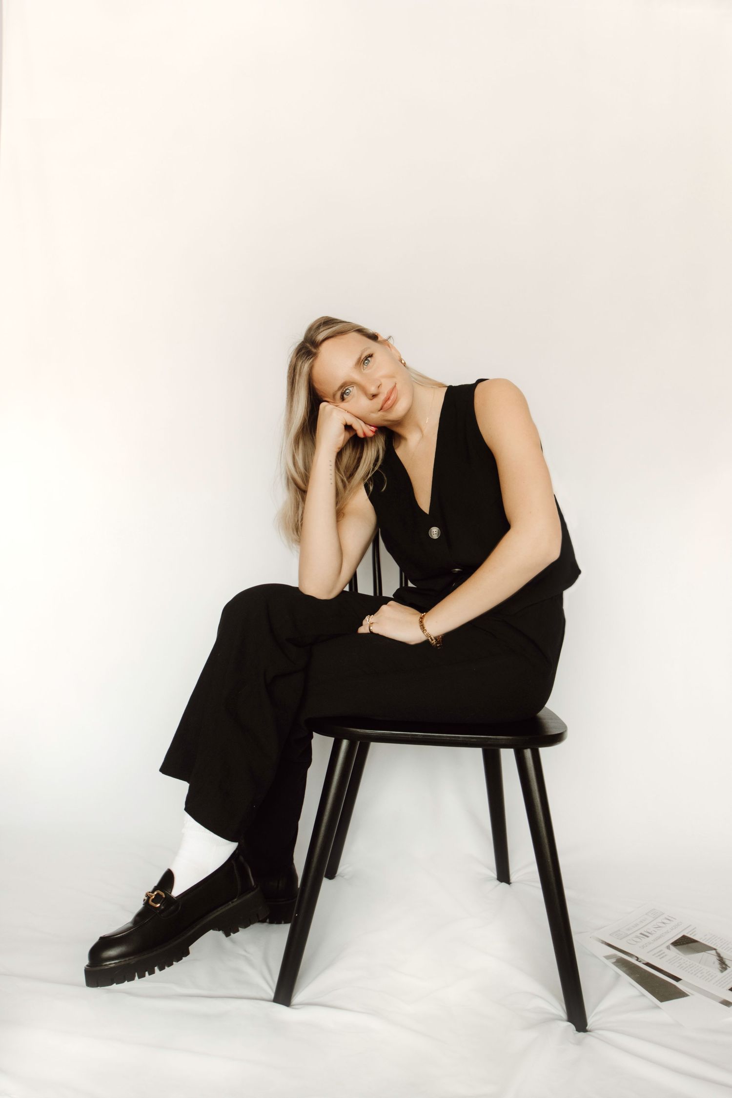 A person in a black jumpsuit and chunky boots poses casually while sitting on a dark wooden chair against a white backdrop.
