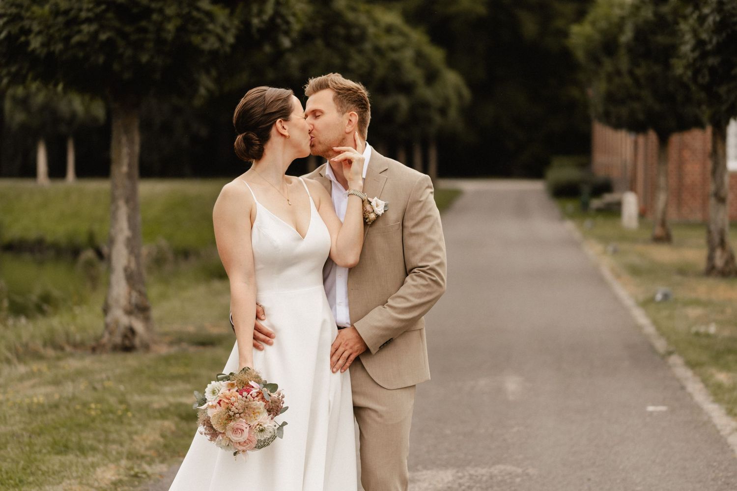 A couple in wedding attire share an intimate moment on a tree-lined path while the bride holds a blush-colored bouquet.