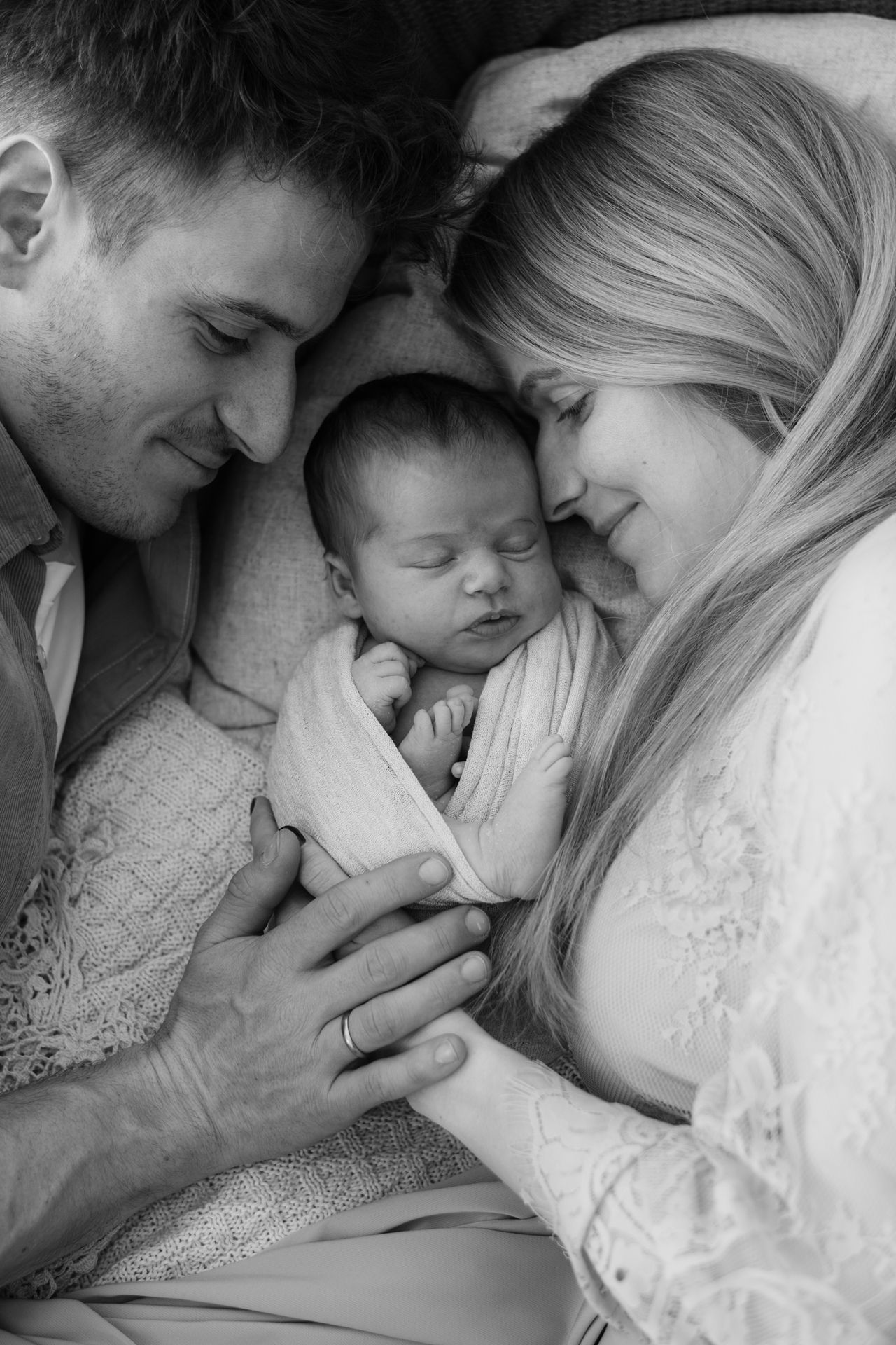Black and white intimate portrait of a couple looking lovingly at their sleeping newborn baby wrapped in blankets.