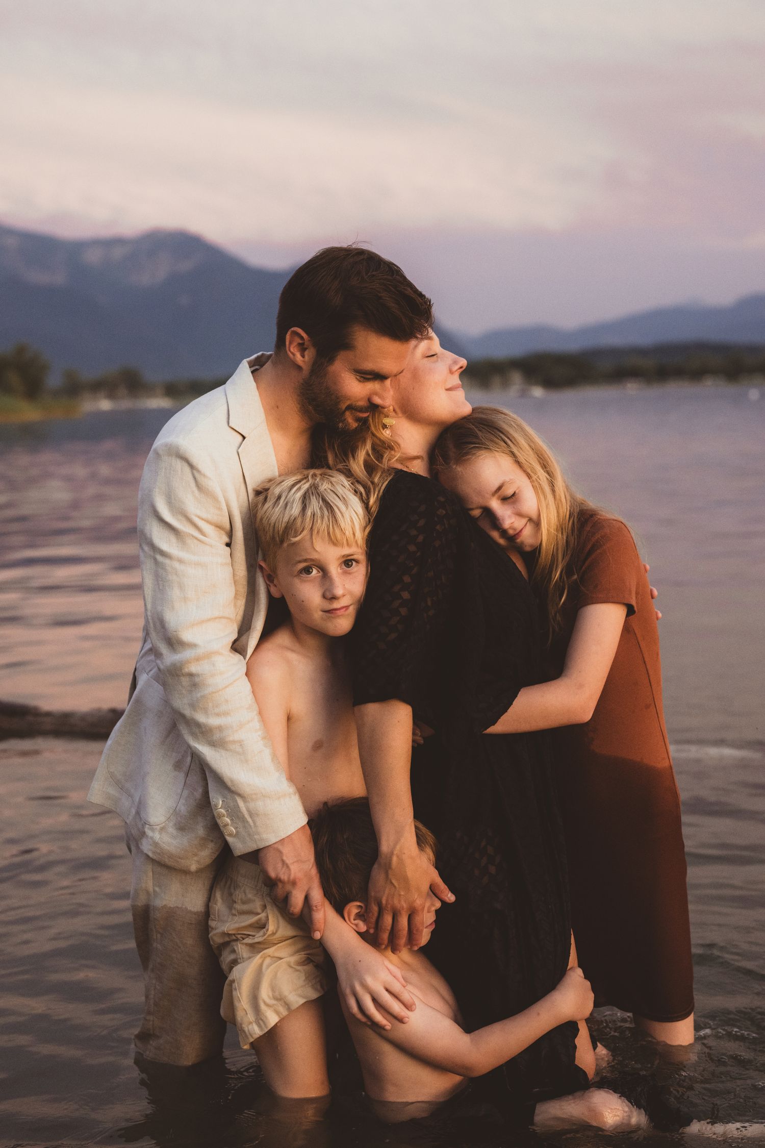 A romantic couple shares intimate moments while standing in shallow water at sunset with mountains in background.