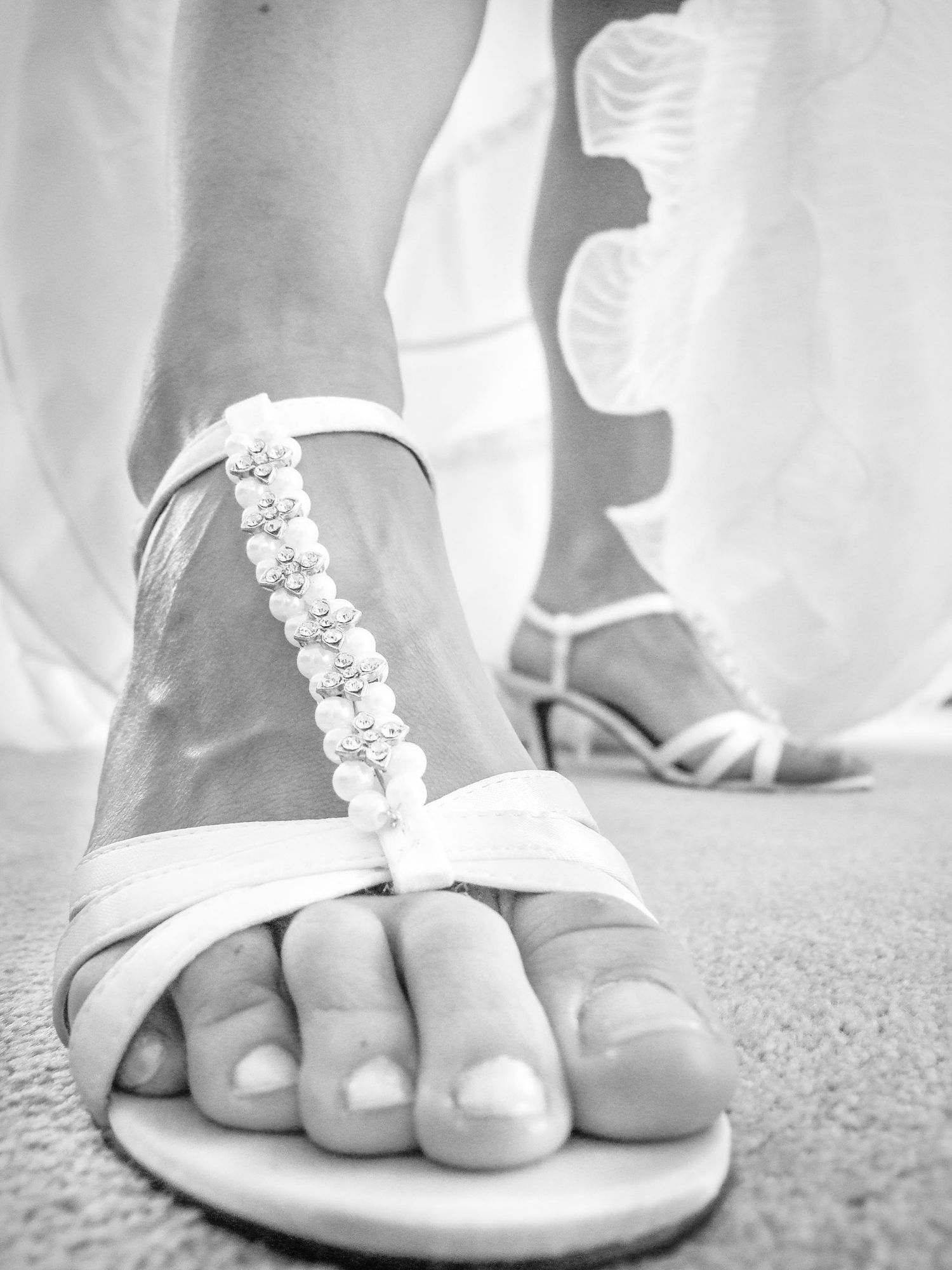 Gros plan en noir et blanc sur les pieds d'une mariée portant des sandales blanches ornées de perles, par Jérémie Botte.