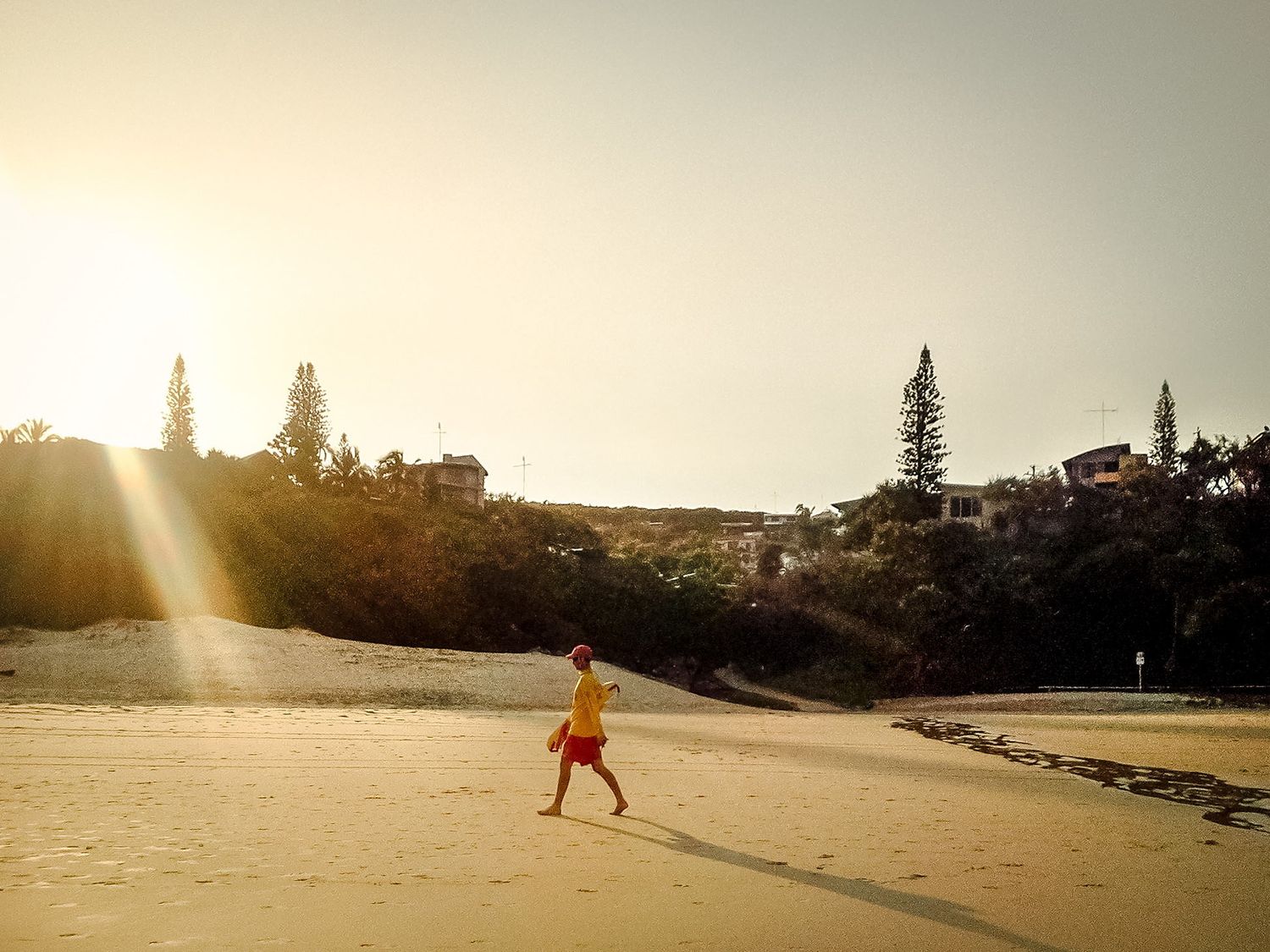 Une personne en short rouge marche sur une grande plage déserte sous la lueur d'un soleil couchant par Jérémie Botte.