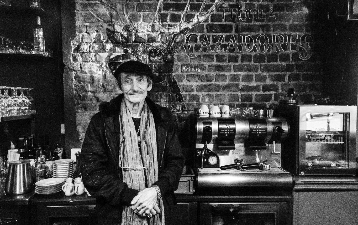 Portrait en noir et blanc d'un homme moustachu en casquette derrière le comptoir d'un bistrot par Jérémie Botte.