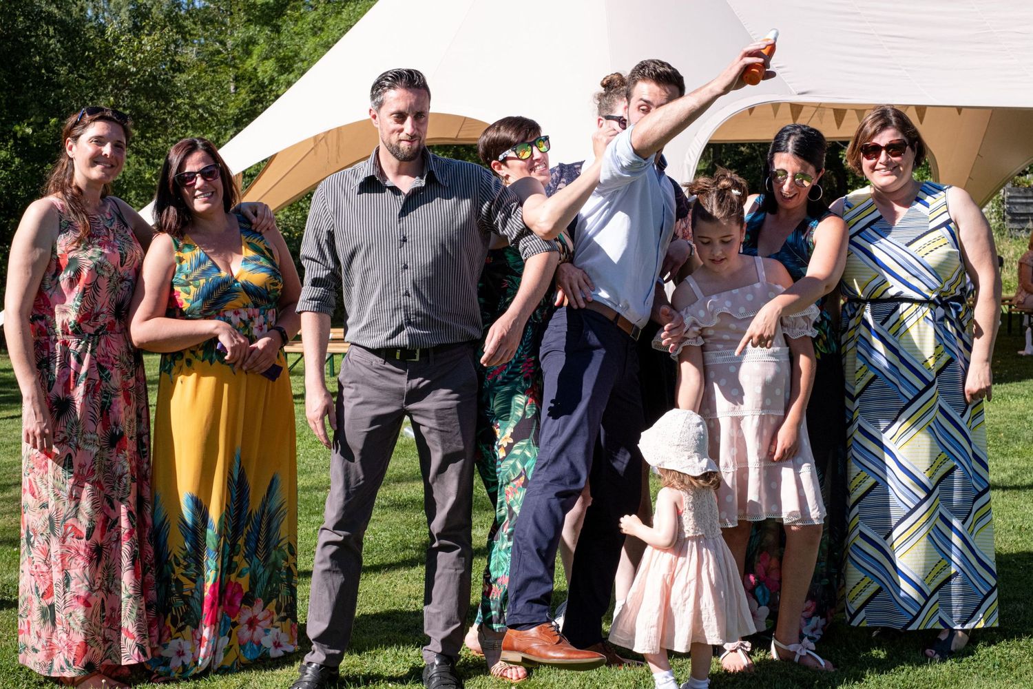 Une joyeuse photo de groupe réunissant adultes et enfants en habits de fête sur une pelouse devant une grande tente, par Jérémie Botte.