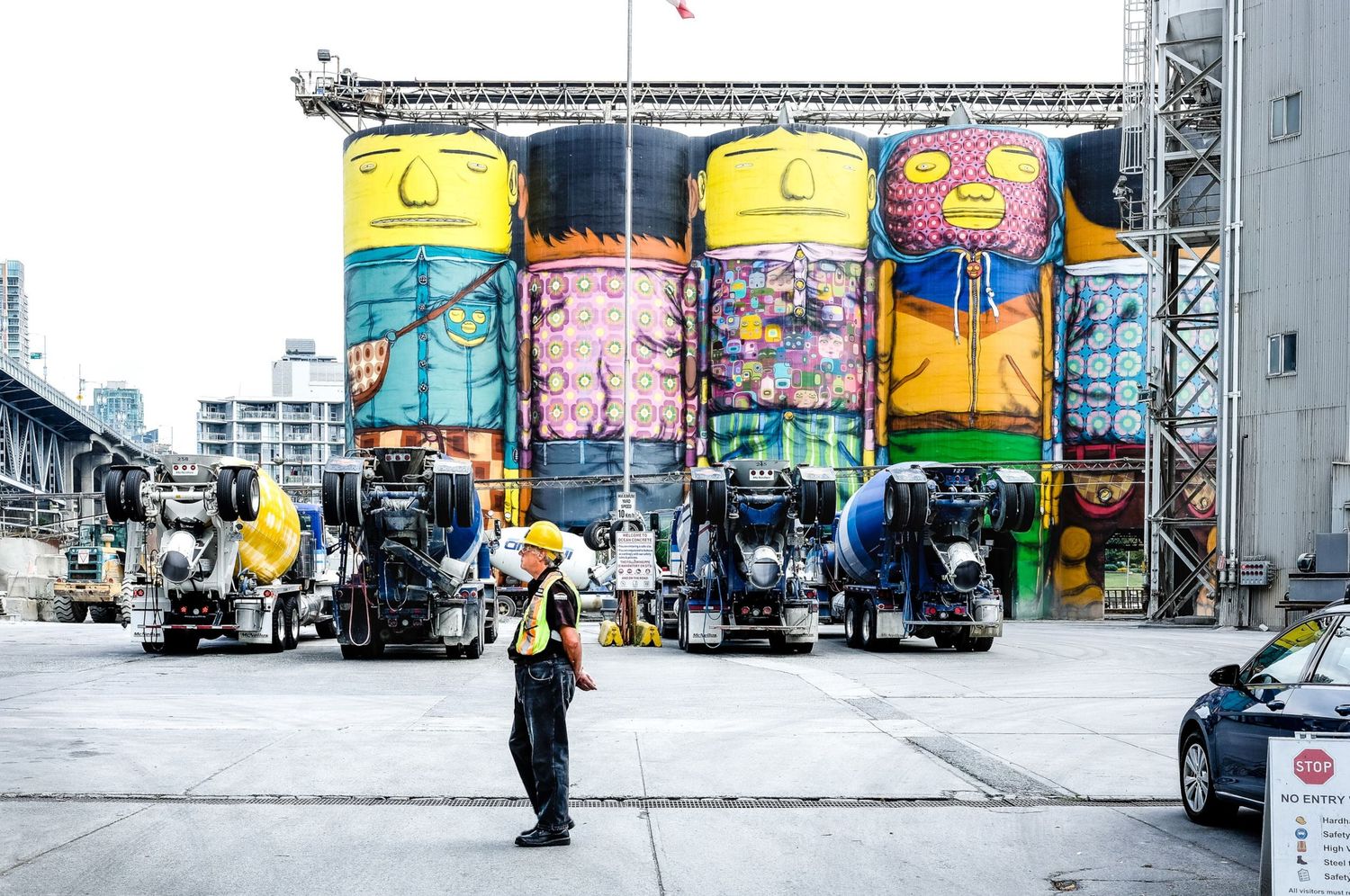 Un ouvrier en gilet de sécurité devant d'immenses silos industriels peints de fresques colorées par Jérémie Botte.