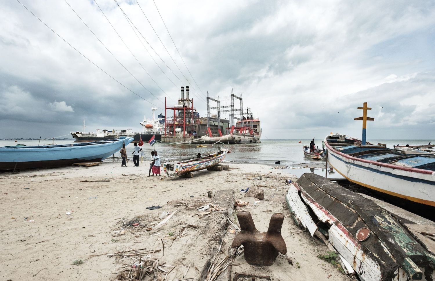 Des barques de pêcheurs traditionnelles posées sur une plage de sable fin devant un complexe industriel en Afrique, par Jérémie Botte.