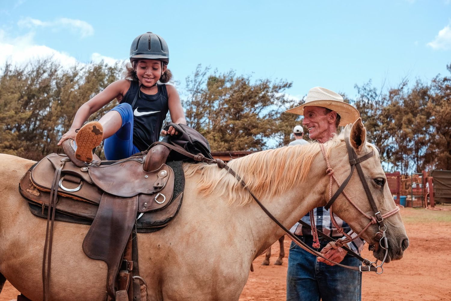 Une jeune fille casquée s'apprête à monter à cheval tenu par un homme au chapeau de cowboy, par Jérémie Botte.