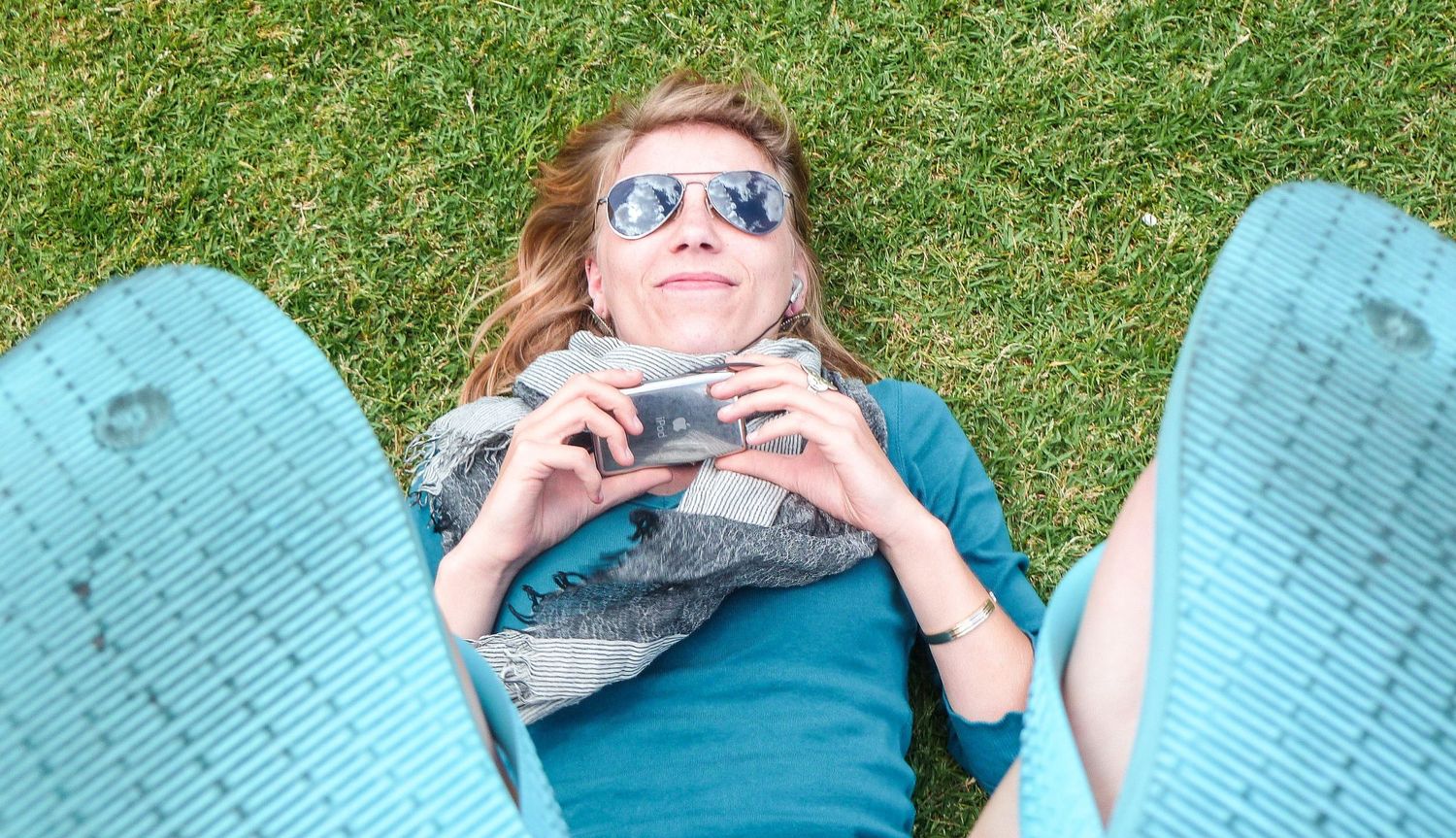 Une femme souriante avec des lunettes de soleil est allongée dans l'herbe, photographiée depuis ses pieds par Jérémie Botte.
