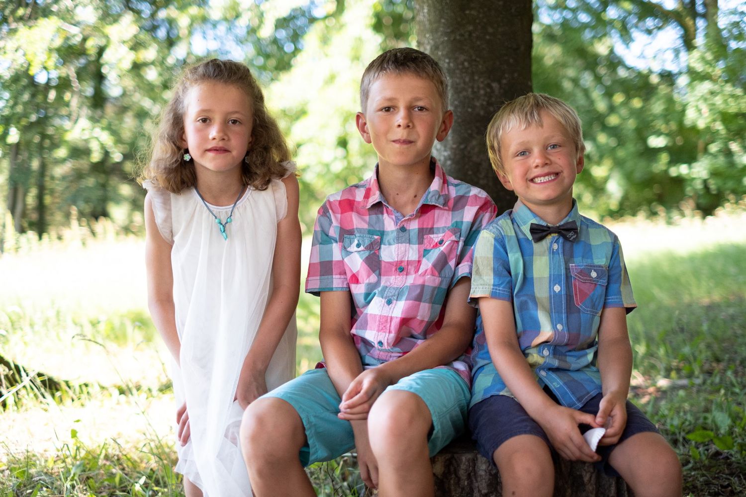 Trois enfants en tenue de fête, une fille et deux garçons, assis sur une souche dans les bois, par Jérémie Botte.