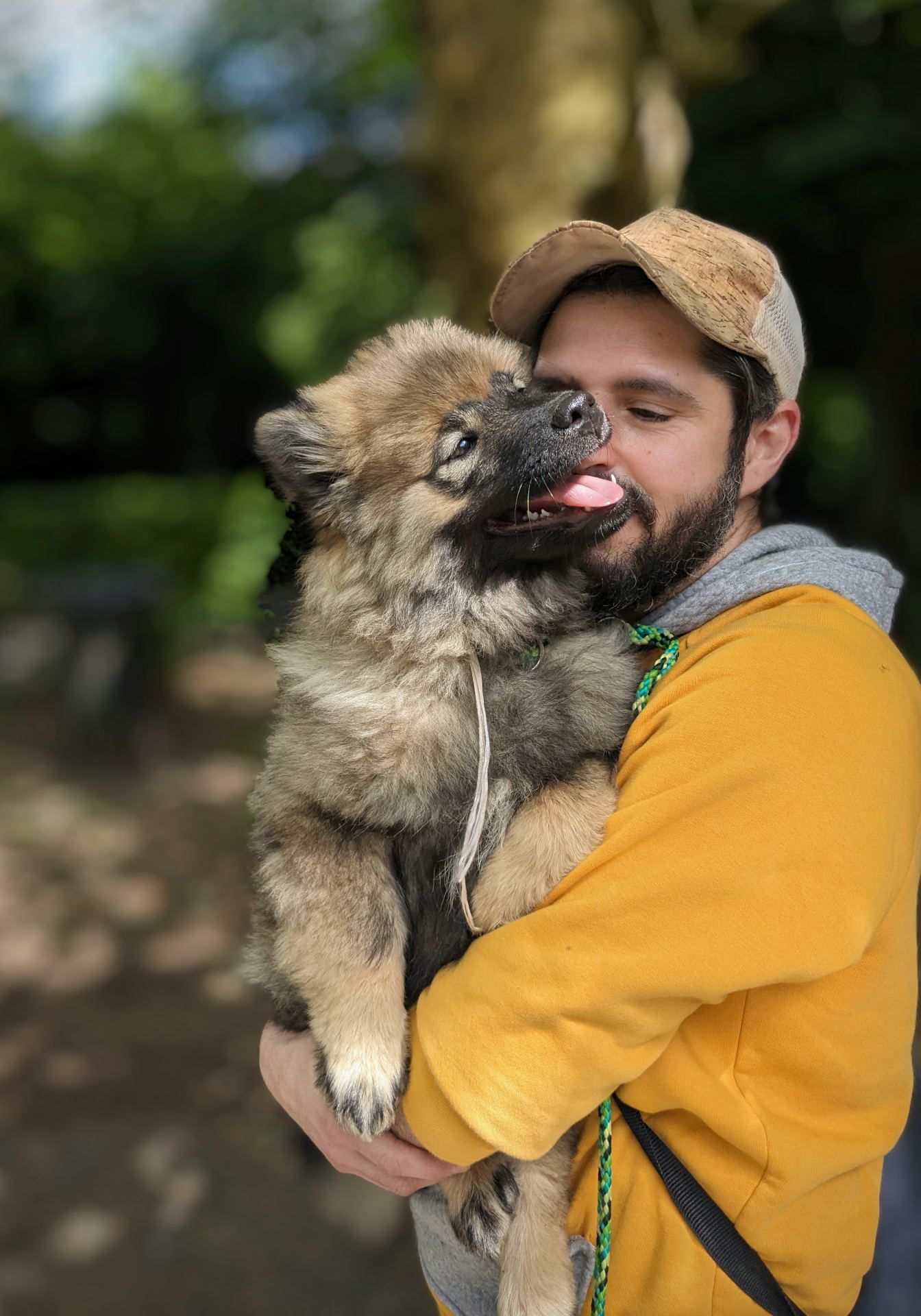 Un homme barbu portant un sweat-shirt jaune et une casquette enlace tendrement un jeune chien eurasier, pour le Portefolio de Jérémie Botte.