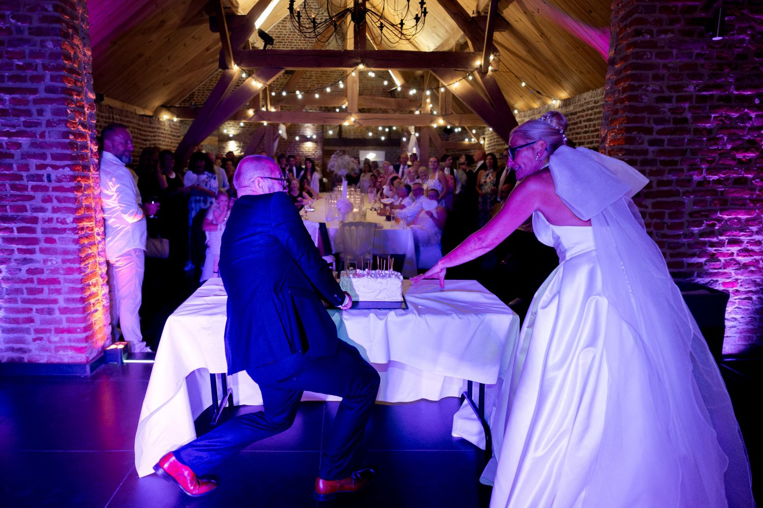 Photographie de mariage d'interieur de nuit de deux mariés découpant un gâteau devant leurs invités par le photographe Jérémie Botte.