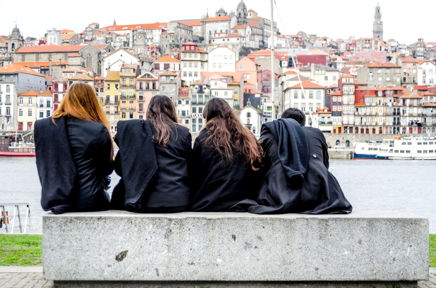 Quatre étudiants portant la cape traditionnelle assis de dos face aux façades colorées de la ville de Porto au Portugal, par Jérémie Botte.