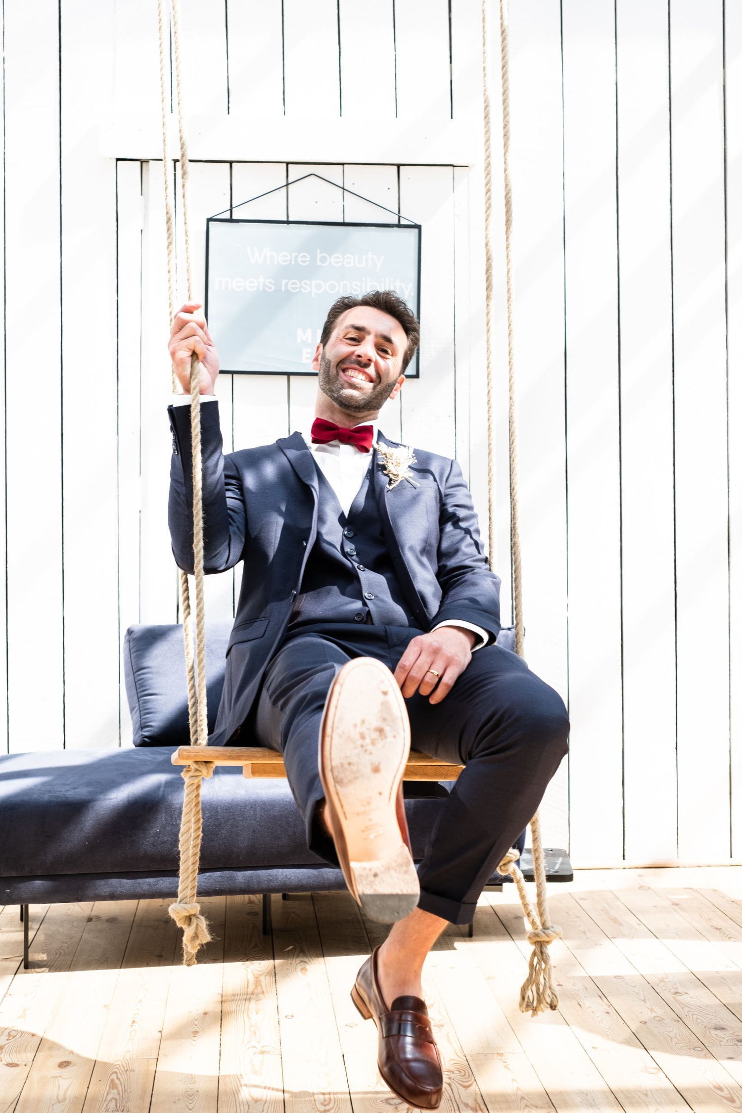 Portrait amusant du marié en costume bleu et nœud papillon rouge, assis tout sourire sur une balançoire en bois, par Jérémie Botte.
